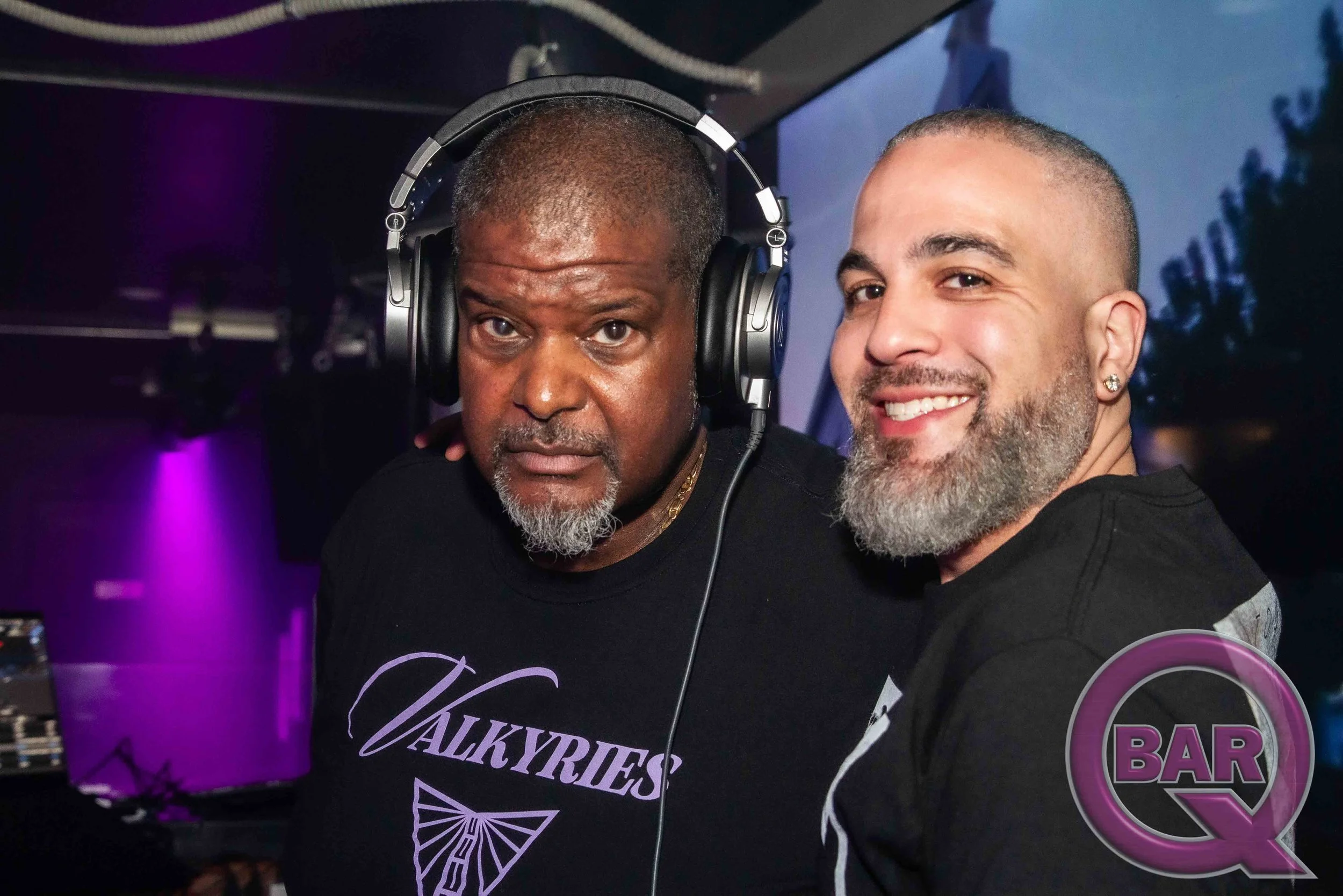 Two men posing for a photo at a bar with purple lighting. One man is wearing headphones and a black shirt with the words 'Walky' and 'Fies' on it. The other man has a beard, earrings, and is smiling.