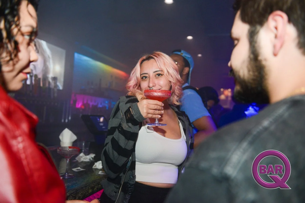 A group of young people socializing at a bar or nightclub. A woman with pink hair and a septum piercing is holding a fancy cocktail glass with a red drink, smiling at the camera. Other people are visible in the background with colorful lighting.