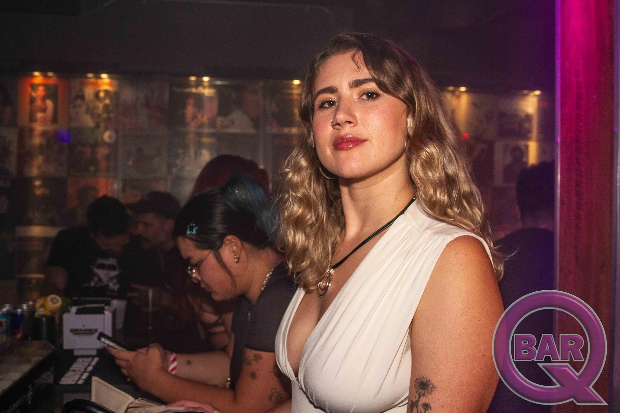 A woman with curly blonde hair and tattoos on her arm standing at a bar in a nightclub, with a colorful, blurred background.
