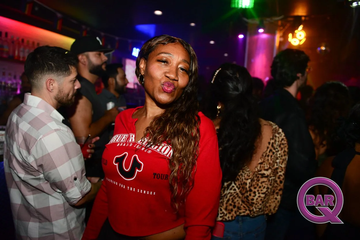 A woman with long wavy hair, wearing a red shirt with a logo and text, making a kissing face while posing at a crowded bar or nightclub with colorful lights and other party-goers in the background.
