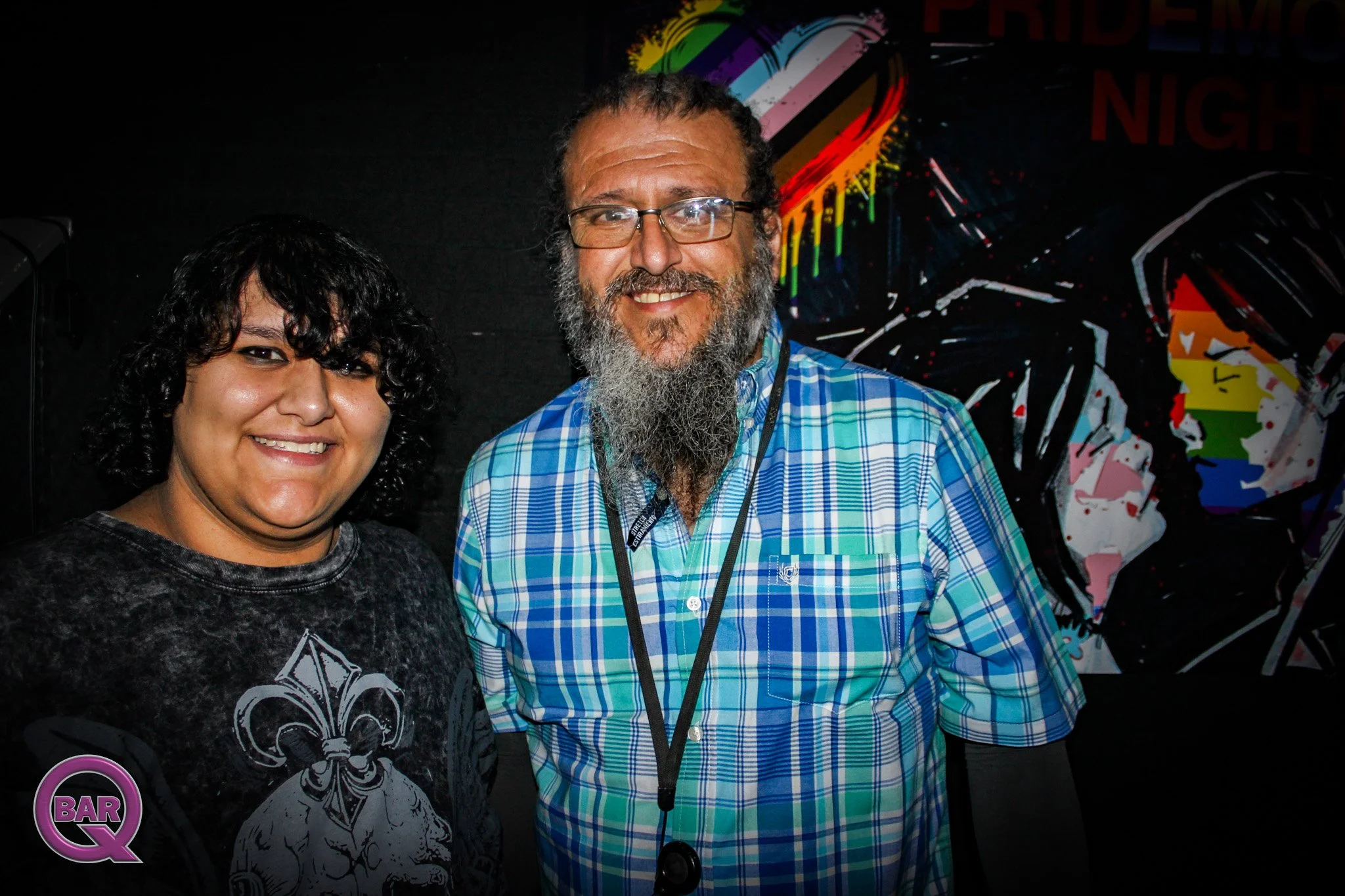 Two smiling people posing at an event, with a colorful rainbow-colored flag or artwork in the background. One person is a woman with dark, curly hair wearing a black shirt with a white design, and the other is a man with glasses, a long beard, and we