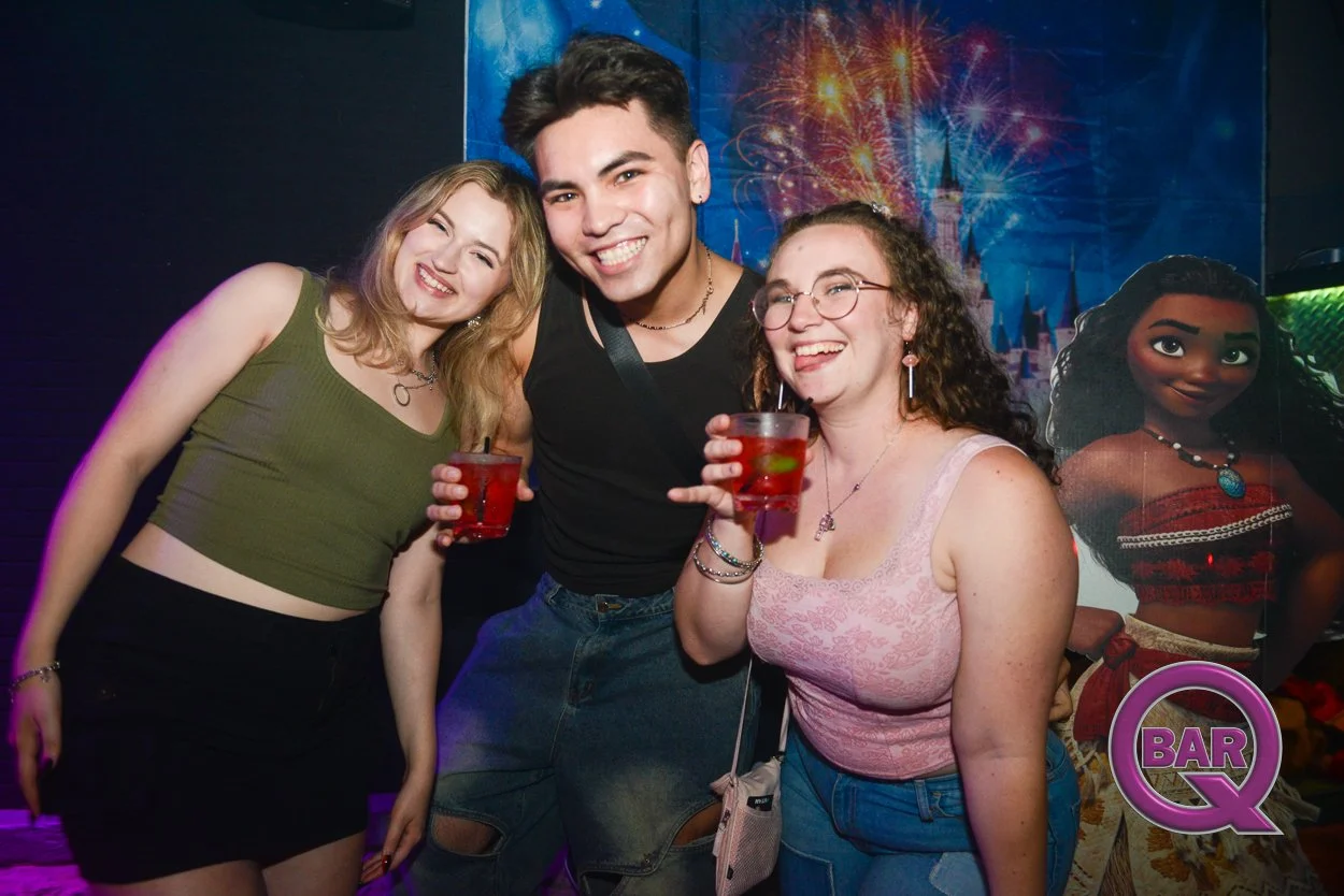 Three young adults smiling and holding drinks at a nightclub with a Disney character poster and firework display background.