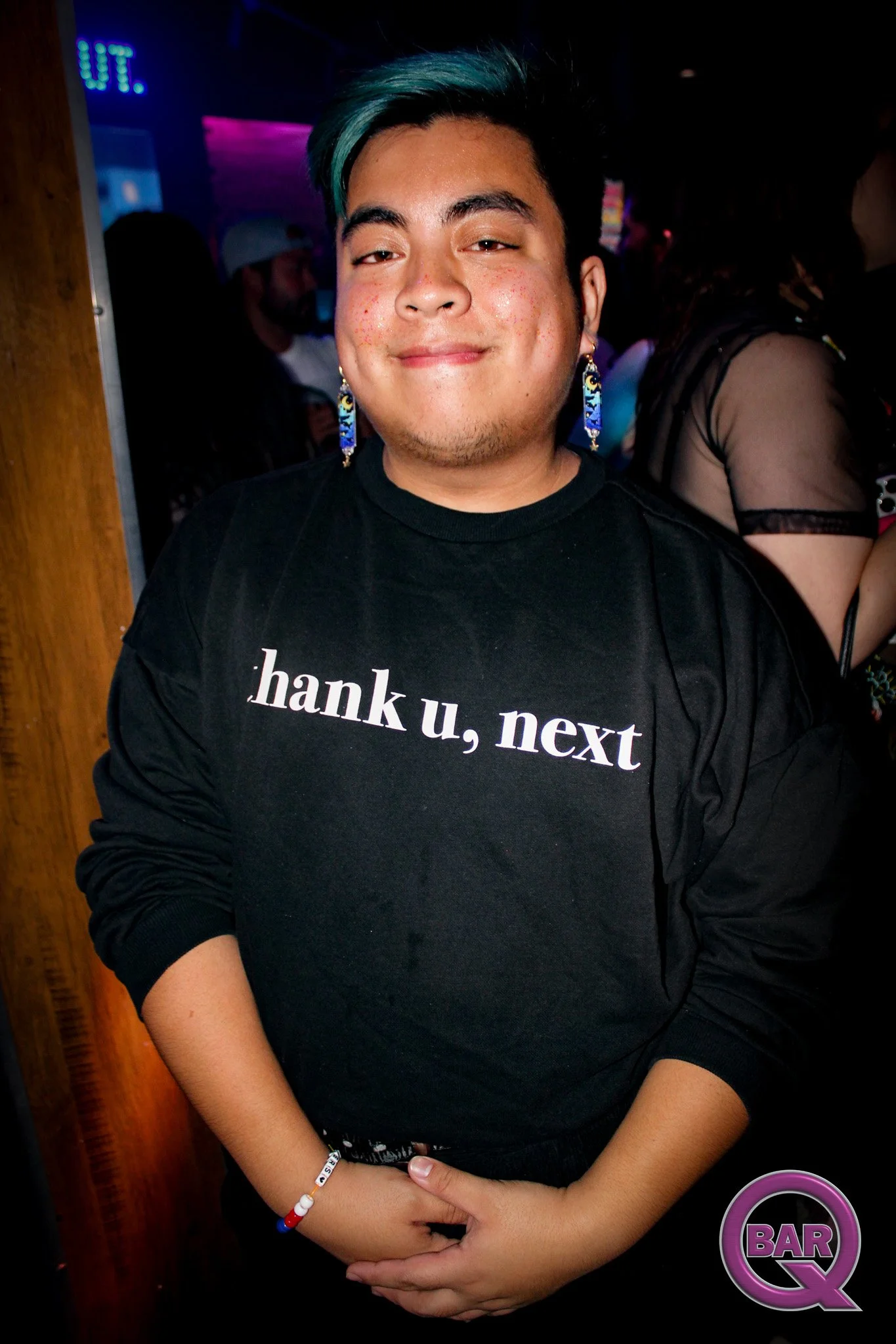 A young person with short dark hair and teal highlights smiling at the camera, wearing a black T-shirt with partially visible white text, colorful earrings, and a beaded bracelet, in a dimly-lit bar or club environment.