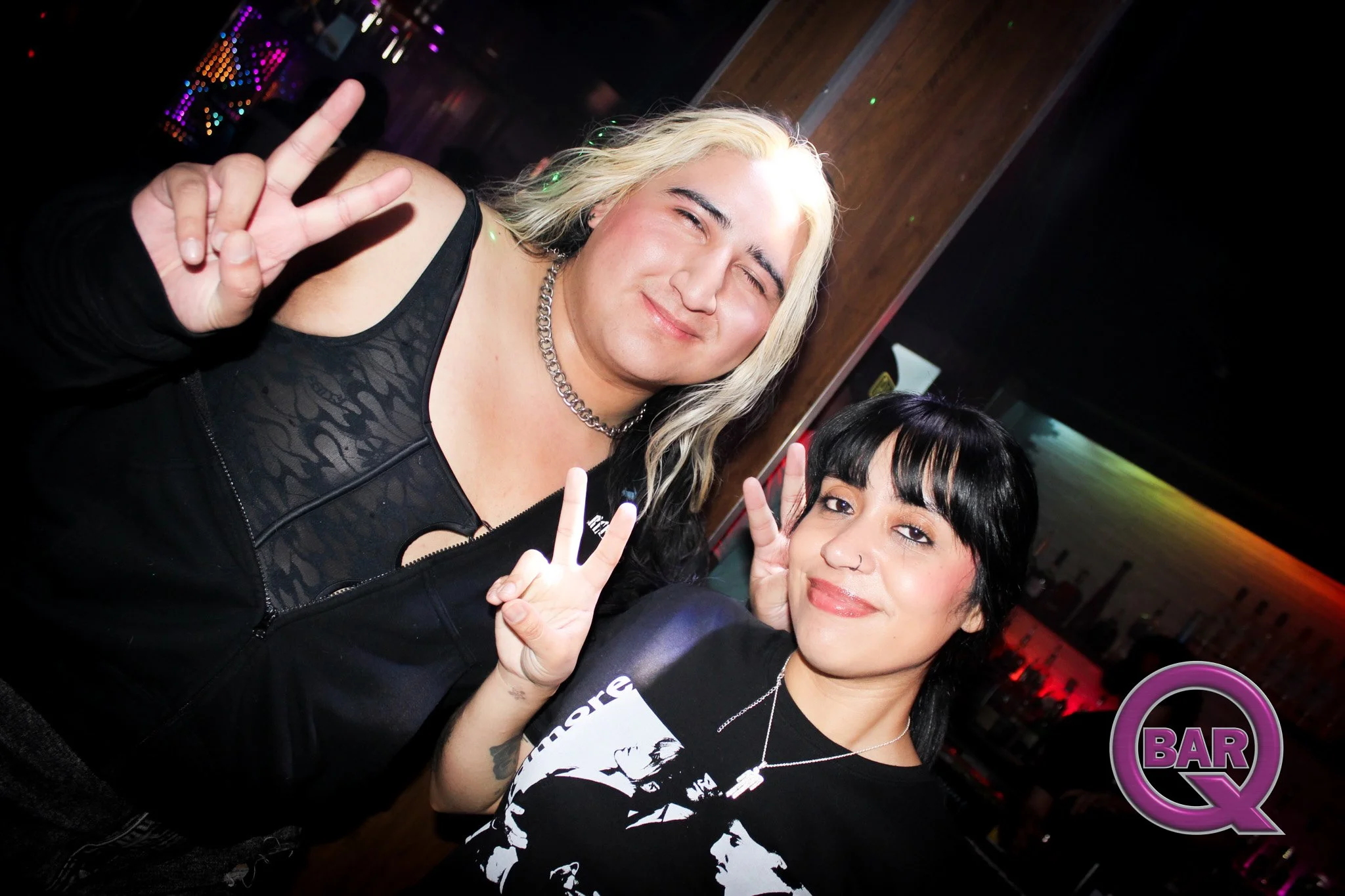 Two women at a bar or nightclub making peace signs with their fingers. One woman has blonde hair and a black outfit, the other has black hair with bangs and a black and white patterned shirt. Colorful lights are in the background.