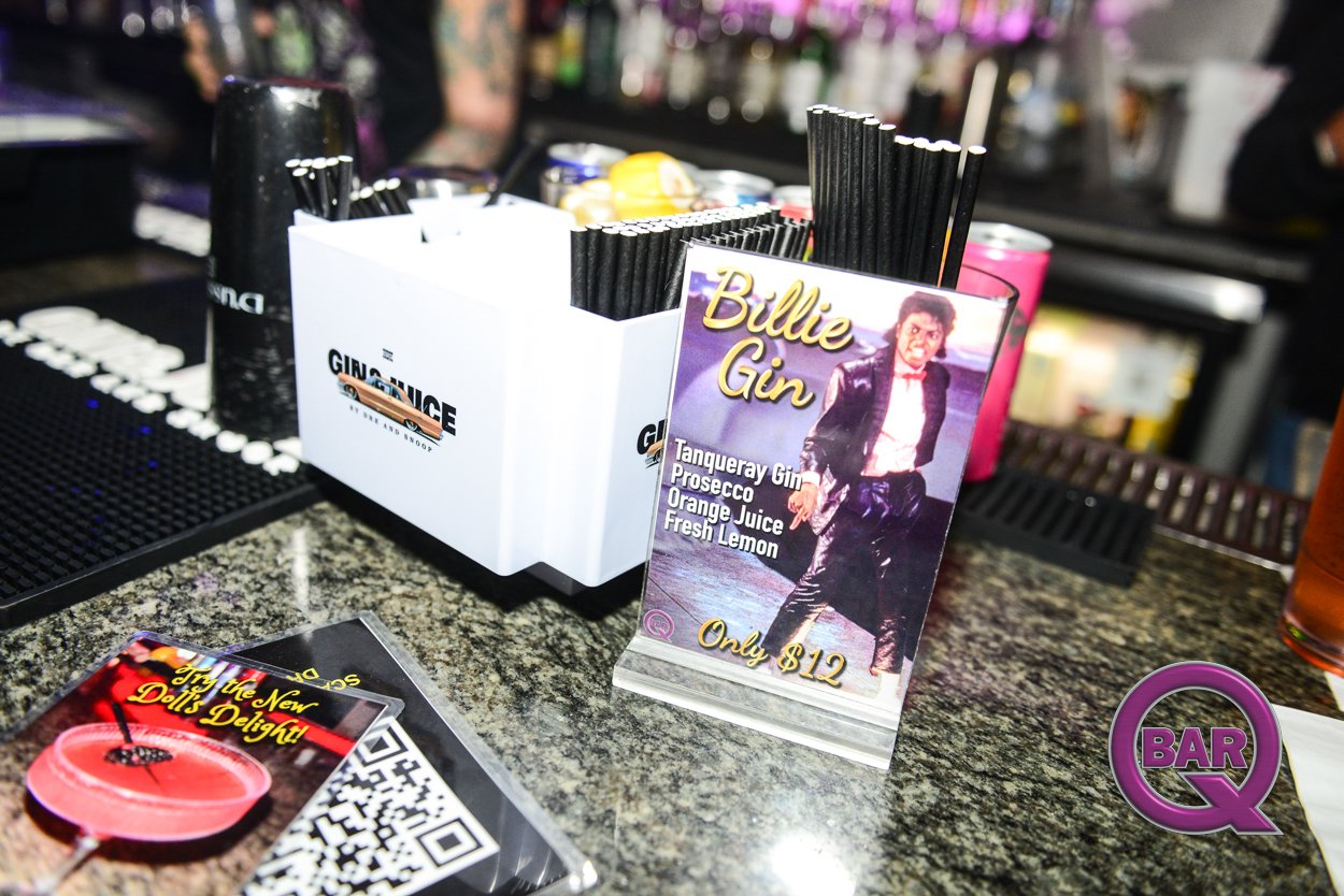 A bar counter with a promotional sign for a cocktail called 'Billie Gin' featuring an image of a woman in a leather jacket, listing ingredients like Tanqueray Gin, Prosecco, orange juice, and fresh lemon, and priced at $12. Various drinks and bar sup