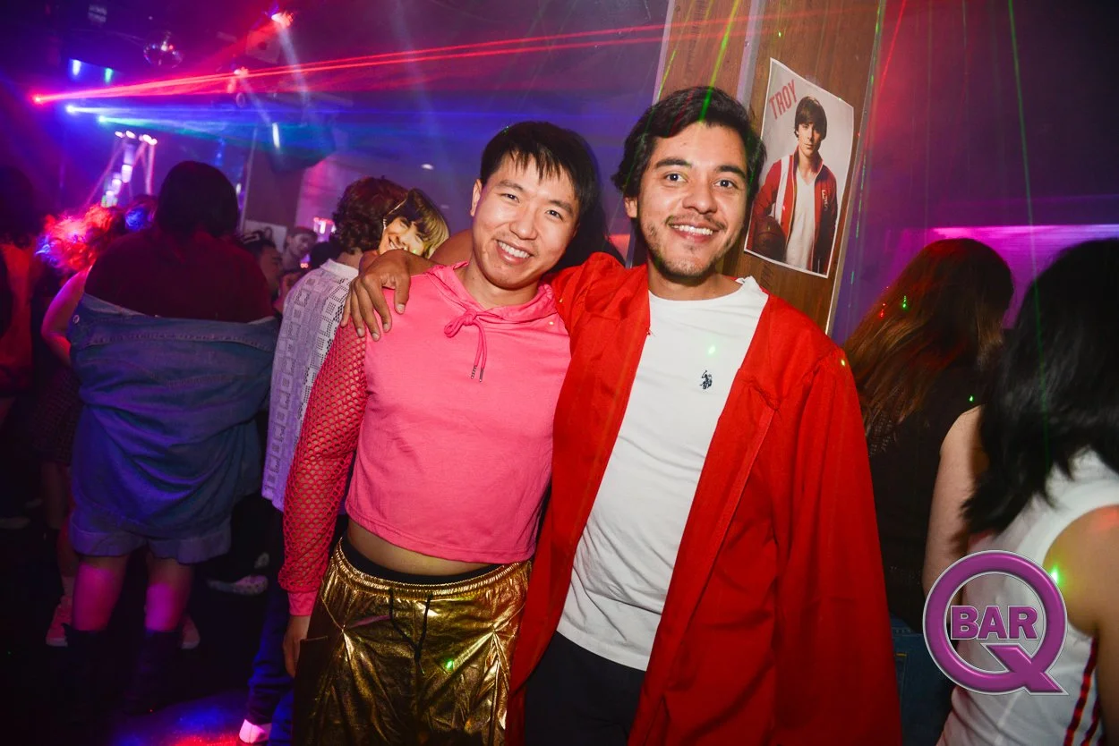 Two smiling men with arms around each other at a nightclub with multicolored laser lights, a wooden wall with a poster of a young man, and a purple BAR Q logo in the lower right corner.