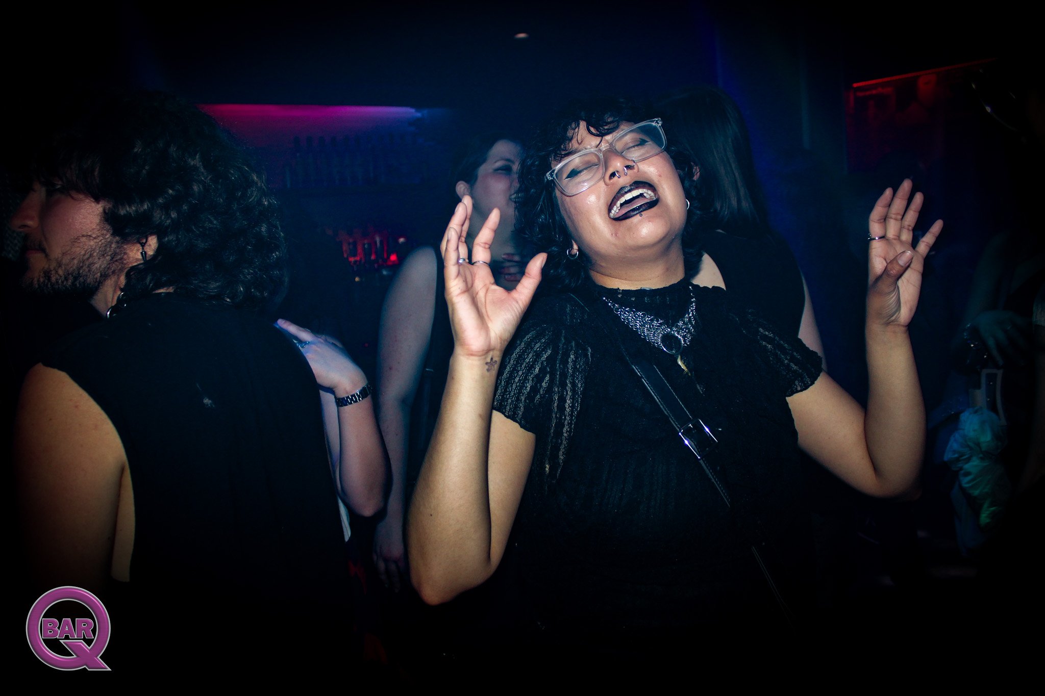 A woman with glasses, curly hair, and a septum piercing enjoys music at a nightclub. Other people dance in the background, with night club lights and a small 'BAR' logo in the corner.