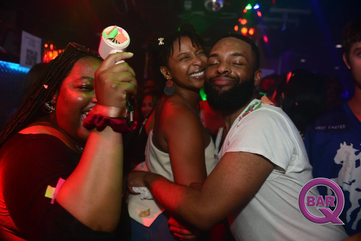 Three people dancing and smiling in a nightclub with colorful lights, one woman holding a small container, and a logo for BAR Q in the bottom right corner.