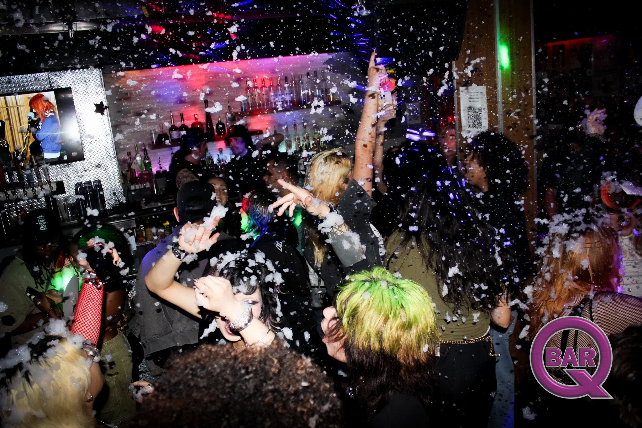 A lively party scene with people dancing, foam, and colorful lights at a bar or nightclub.