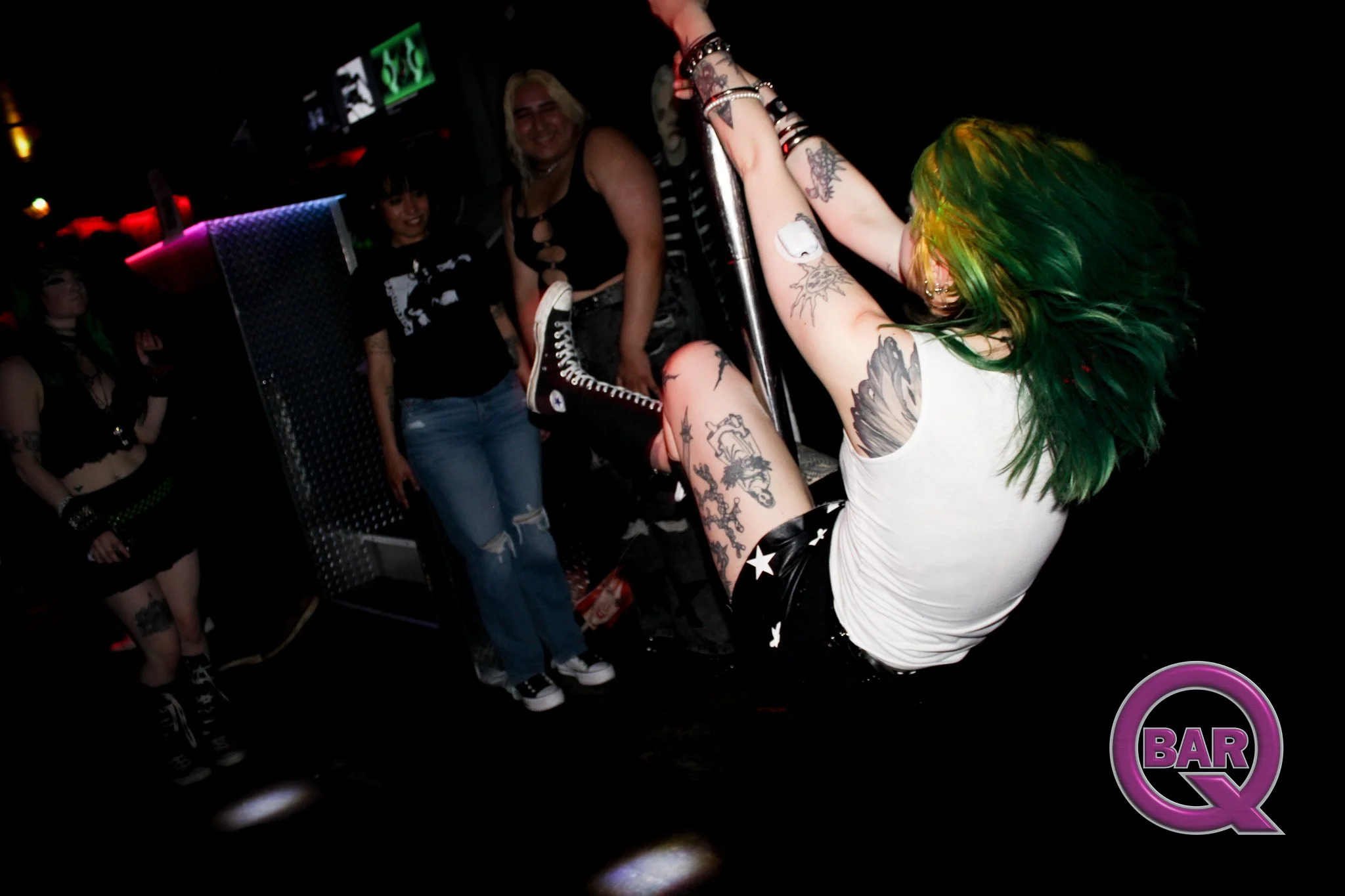 People dancing at a bar with a screen and neon lighting in the background. One woman with green hair and tattoos is on a pole, wearing a white top and black shorts with stars. Other women and a man are nearby, some smiling and dressed in casual or al