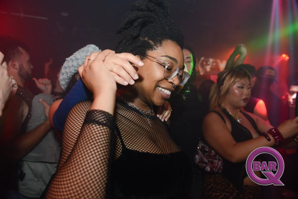 People dancing at a nightclub with colorful lights, including a woman in the foreground wearing glasses, a black fishnet top, and a necklace, smiling while dancing.