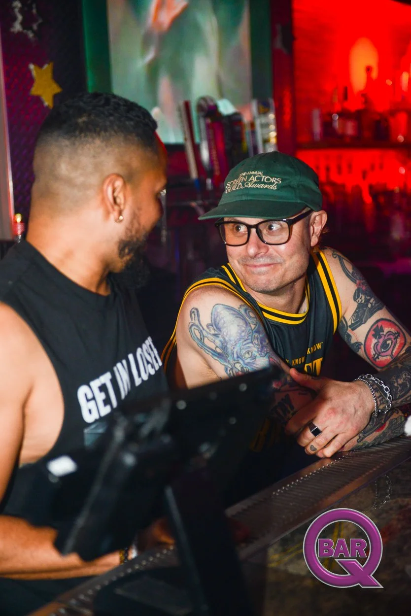 Two men seated at a bar, engaged in conversation. One man has bright tattoos on his arms, wears glasses, a green cap, and a sleeveless sports jersey. The other man wears a black sleeveless shirt with white text. The background features colorful light