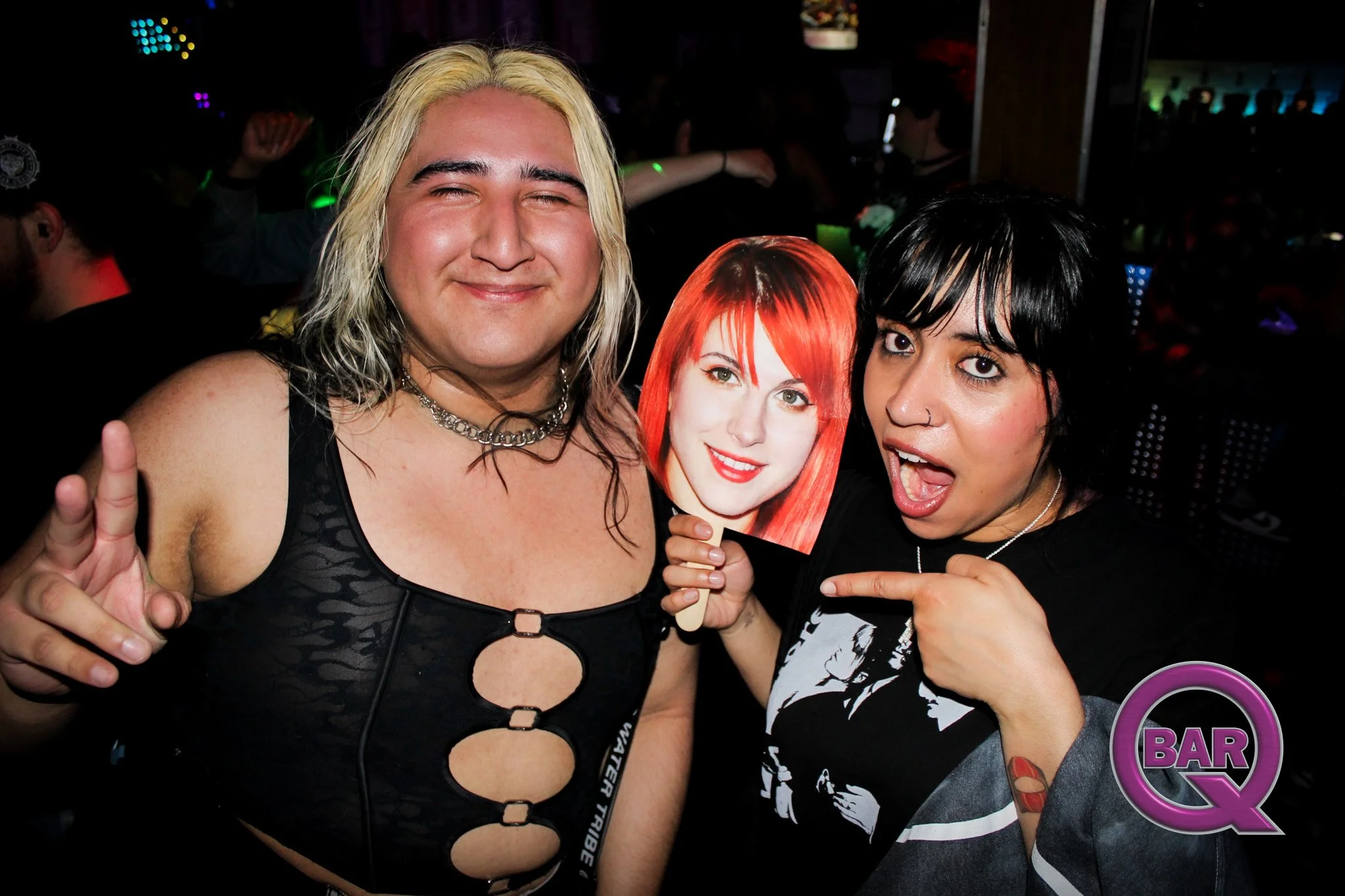 Two women in a nightclub, one with bleached blonde hair and the other with black hair, holding a cardboard cutout of a red-haired woman, with a man in the background making a peace sign gesture.