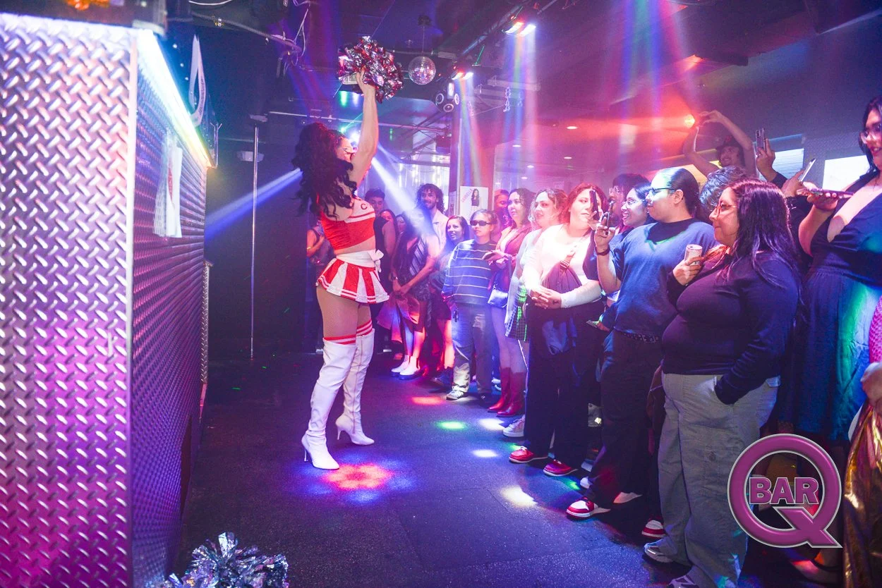 A drag queen performing on stage with an audience watching in a nightclub with colorful lights.