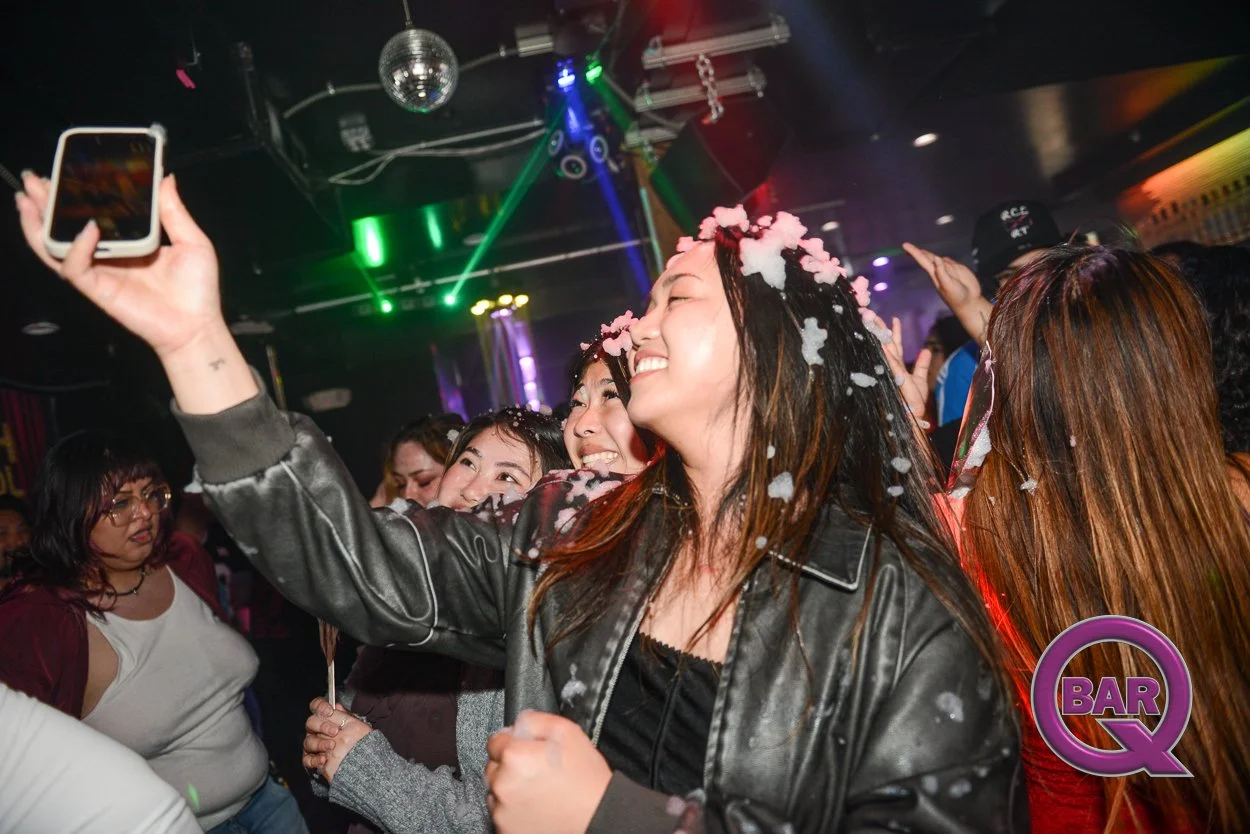 A group of women at a party or nightclub dancing and taking selfies with colorful lights and a disco ball overhead, with a logo that says 'BAR Q' in the bottom right corner.