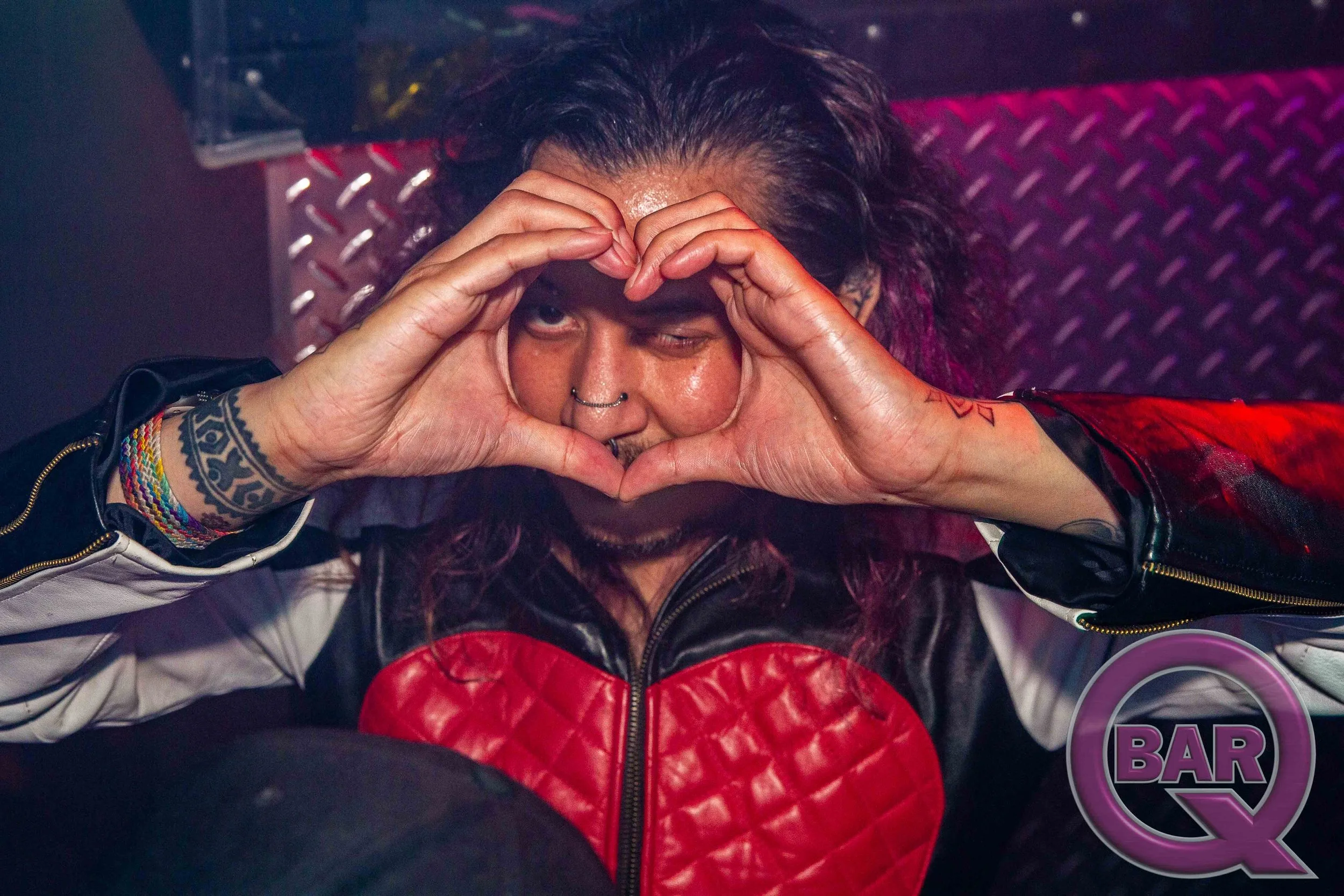 Person with dark hair and tattoos making a heart shape with hands around right eye in a bar setting