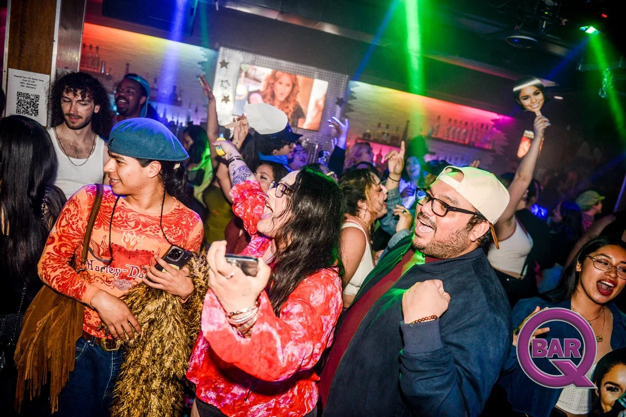 People dancing and socializing at a lively nightclub or bar with colorful lighting, a large screen displaying a woman, and a bartender's shelves in the background.