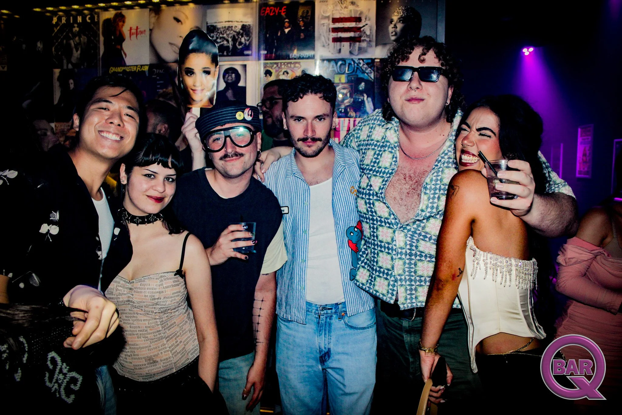 A group of people at a nightclub or bar, standing close together and smiling, with cartoon-style posters of musicians and celebrities on the wall behind them, and purple lighting in the background.