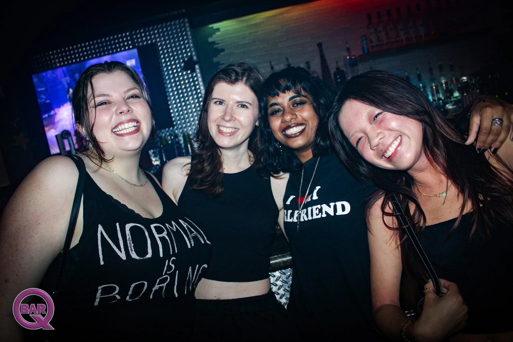 Group of four women smiling and posing in a nightclub or bar with a bar counter and bottles in the background.