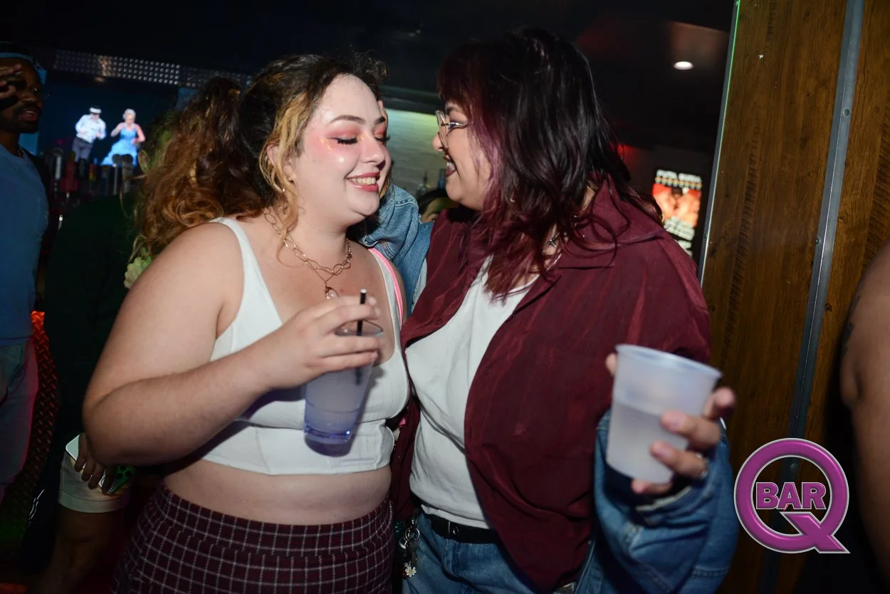 Two women smiling and enjoying drinks together at a bar, engaging in a close conversation. The woman on the left has curly hair and is wearing a white crop top and maroon checkered pants. The woman on the right has shoulder-length dark hair and glass