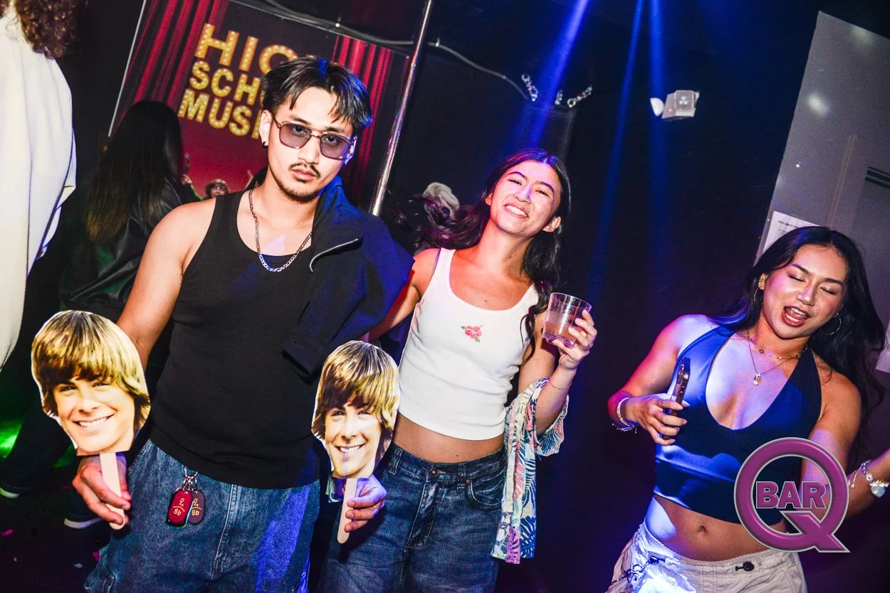 Group of people socializing at a bar, with colorful lighting and a sign that says 'High School Music' in the background. One person holds a drink, and another holds cutouts of a young man's face.