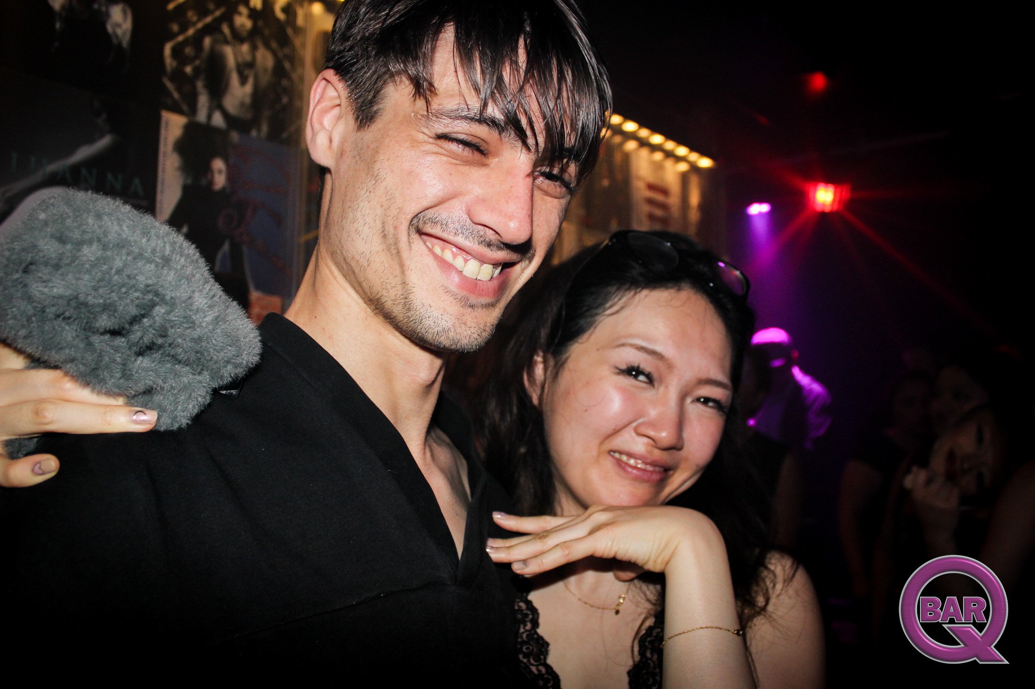 Smiling man with short dark hair and a woman with dark hair in a bar or nightclub with colorful lights and posters in the background.