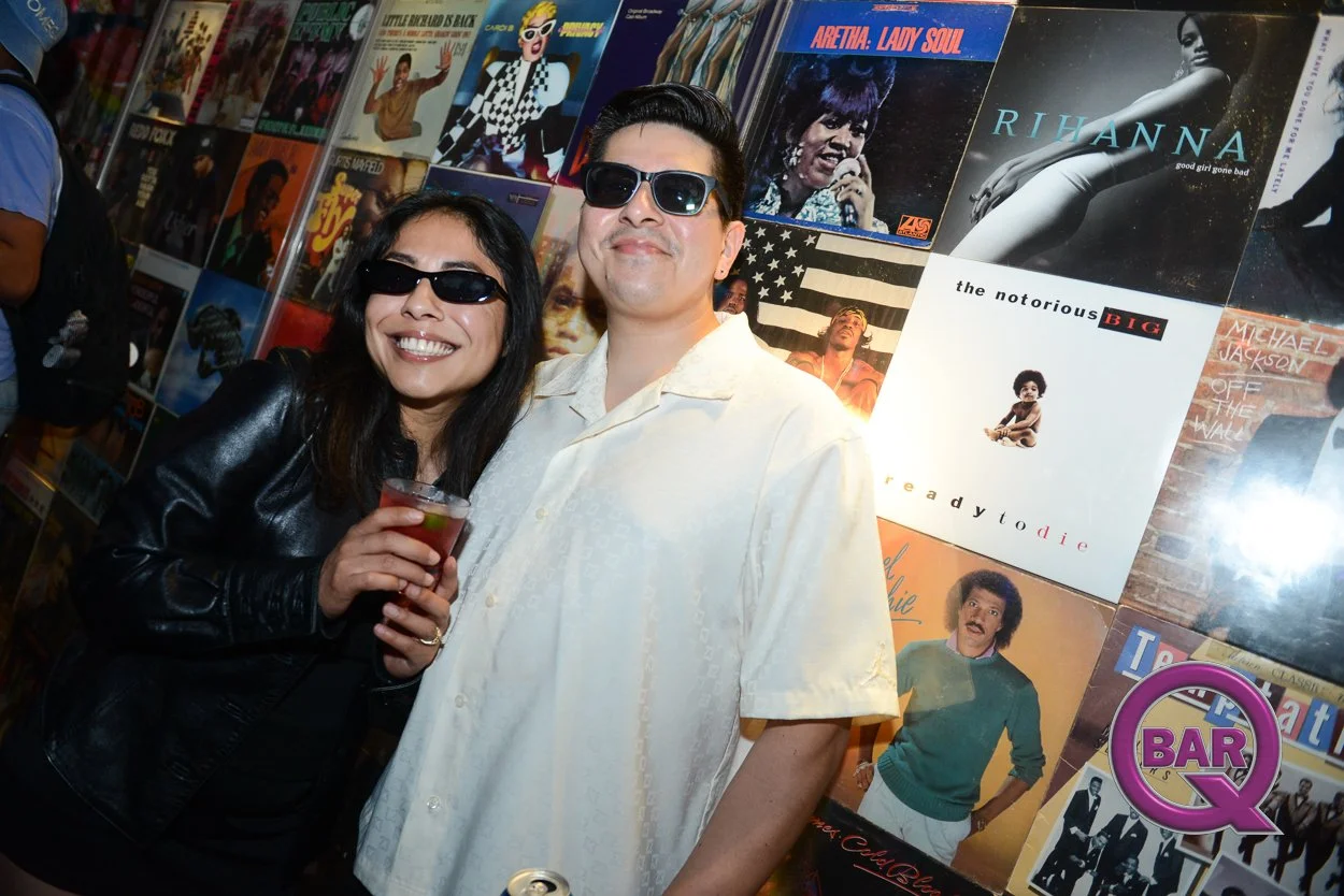 Two people wearing sunglasses standing in front of a wall of vintage vinyl album covers, smiling at an event, with one holding a drink.