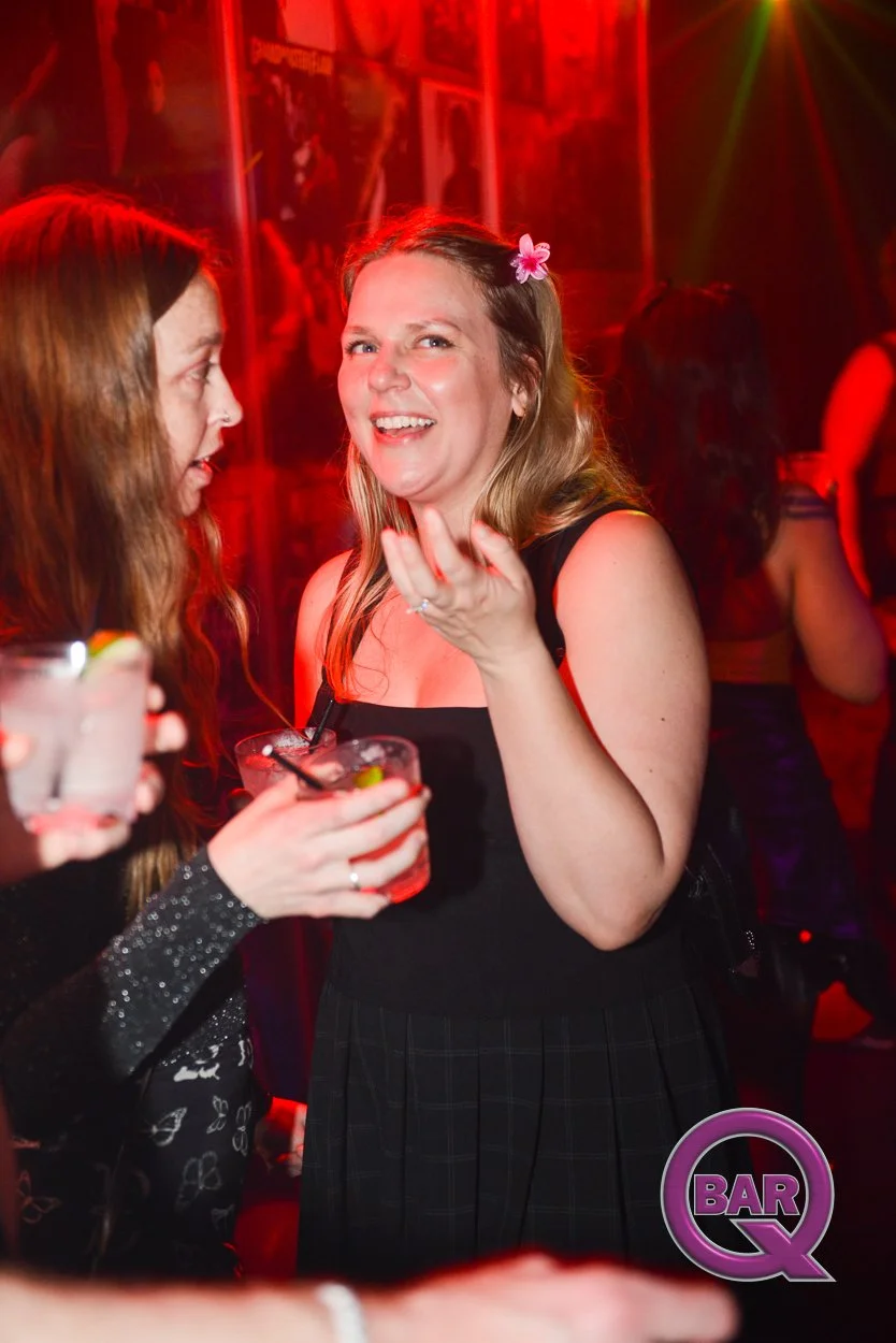 Two women are talking and laughing at a bar or nightclub, with colorful red and green lighting in the background. One woman with long brown hair and a hair flower holds a drink, while the other woman with long reddish-brown hair, wearing a black dres