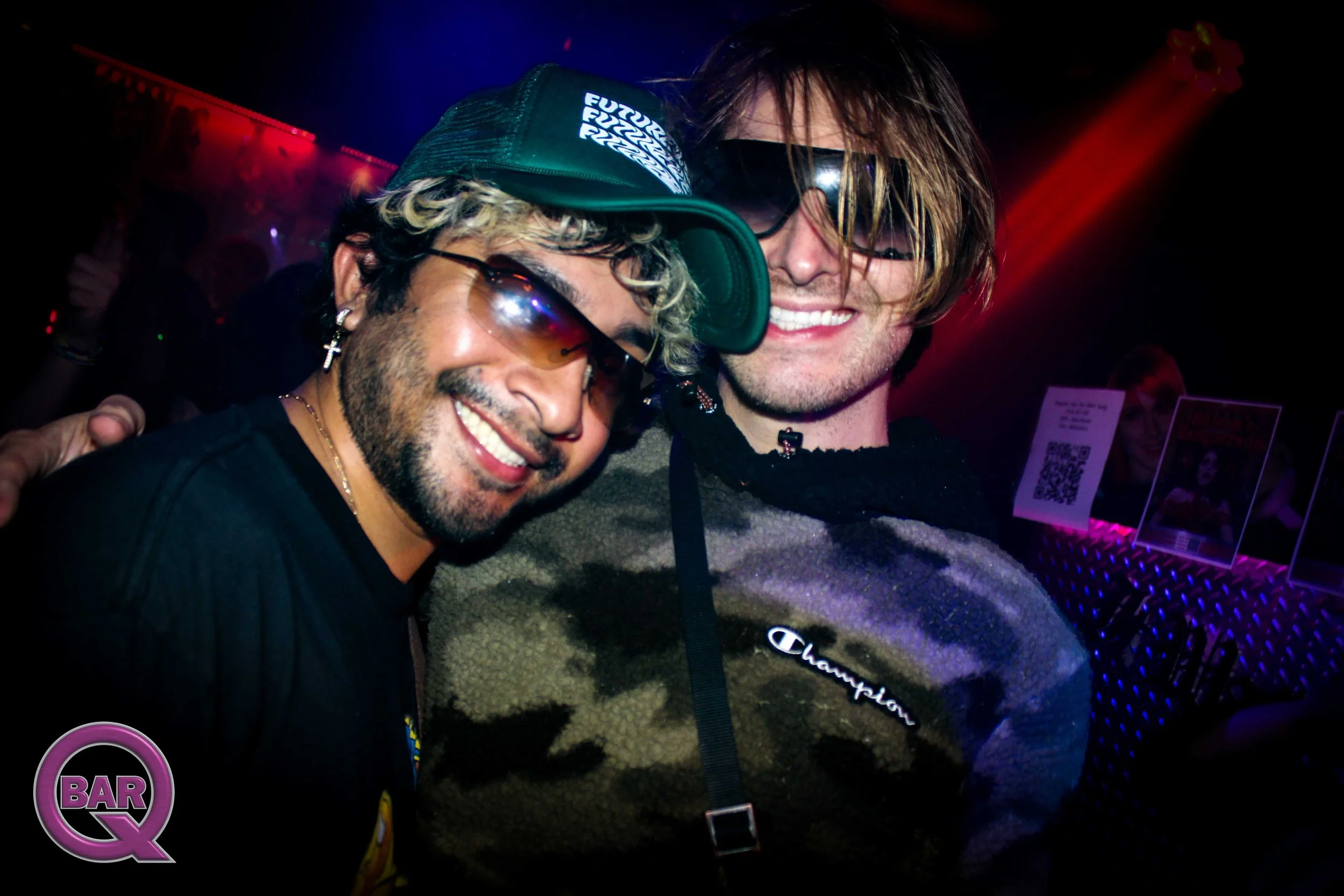 Two men smiling and posing closely together in a dimly lit nightclub or party setting, wearing sunglasses, with one wearing a baseball cap and the other with long hair, surrounded by colorful lights.