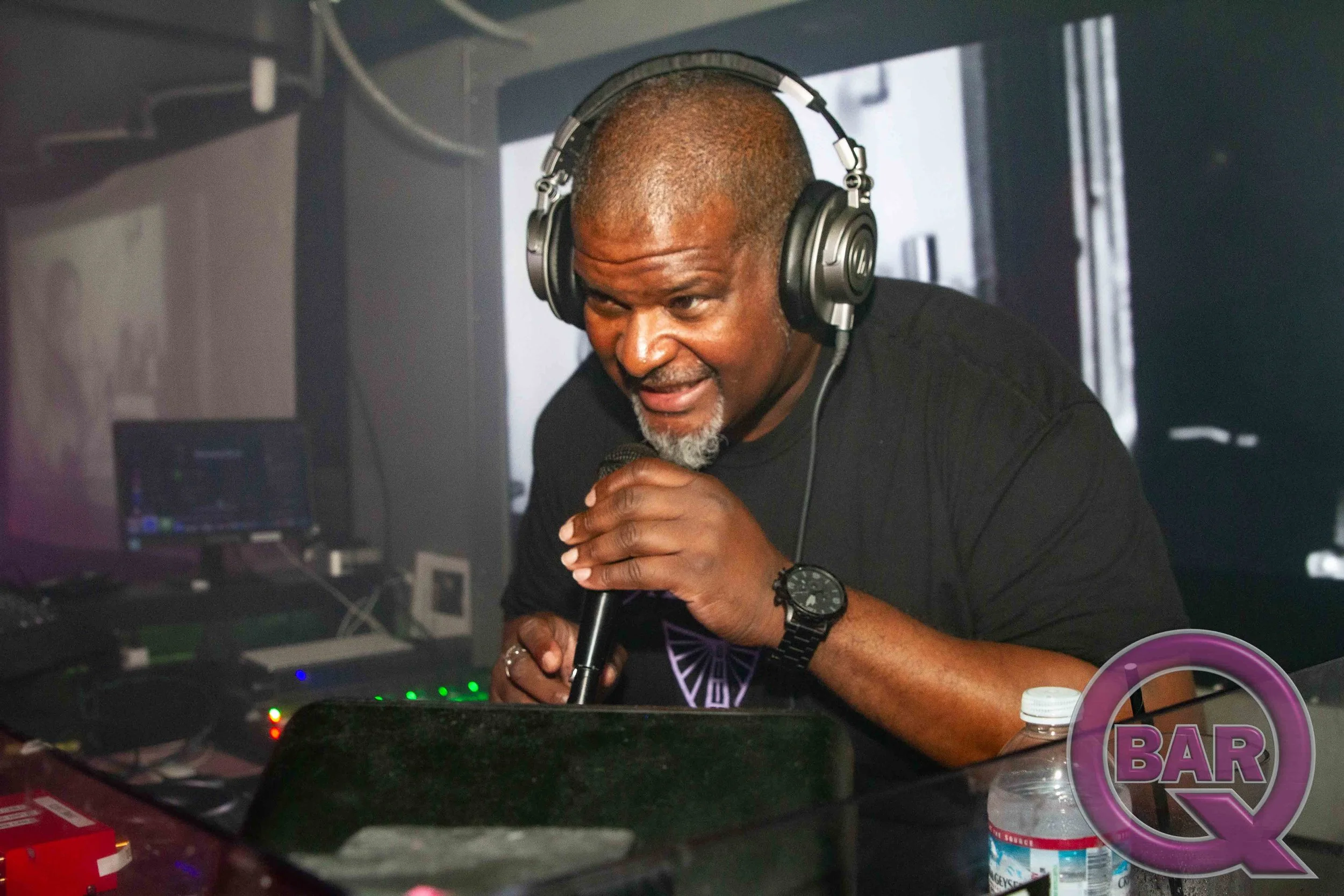 A man with a shaved head and gray beard wearing headphones is speaking into a microphone in a recording studio with computer equipment and bottles on the counter.