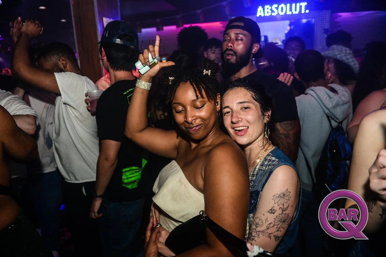 Two women dancing and enjoying themselves at a lively nightclub, surrounded by other partygoers, with a neon sign reading 'ABSOLUT' in the background.