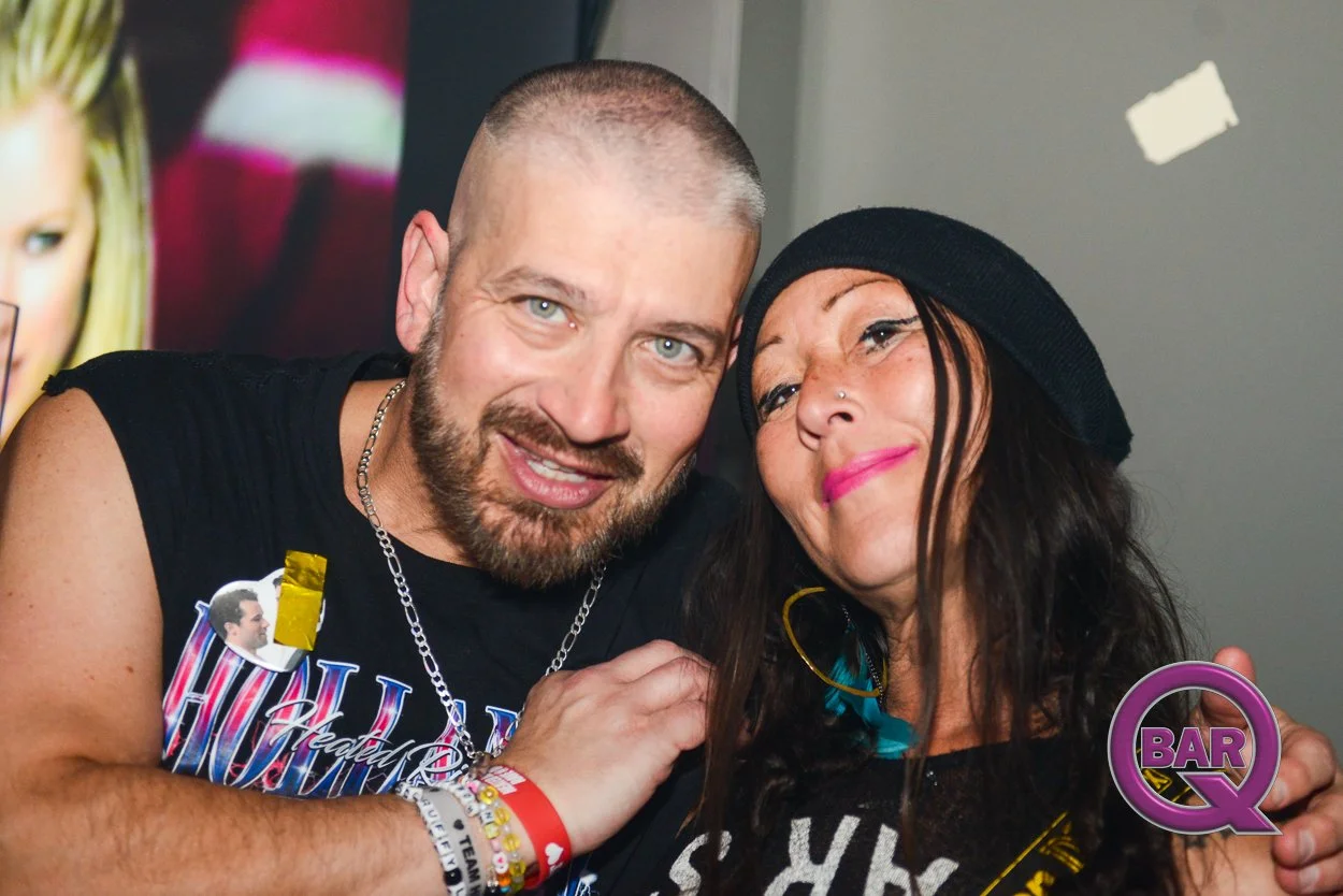 A man and a woman are smiling and leaning close together in a crowded social setting. The man has a beard, short hair, and is wearing a sleeveless black shirt with a colorful design, jewelry, and wristbands. The woman has dark hair, a nose piercing, 