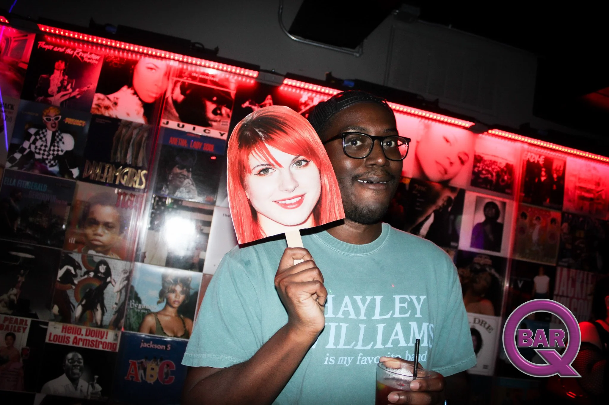 A man holding a cutout of Hayley Williams' face while standing in front of a wall decorated with various posters of musicians and celebrities at a bar or music venue.