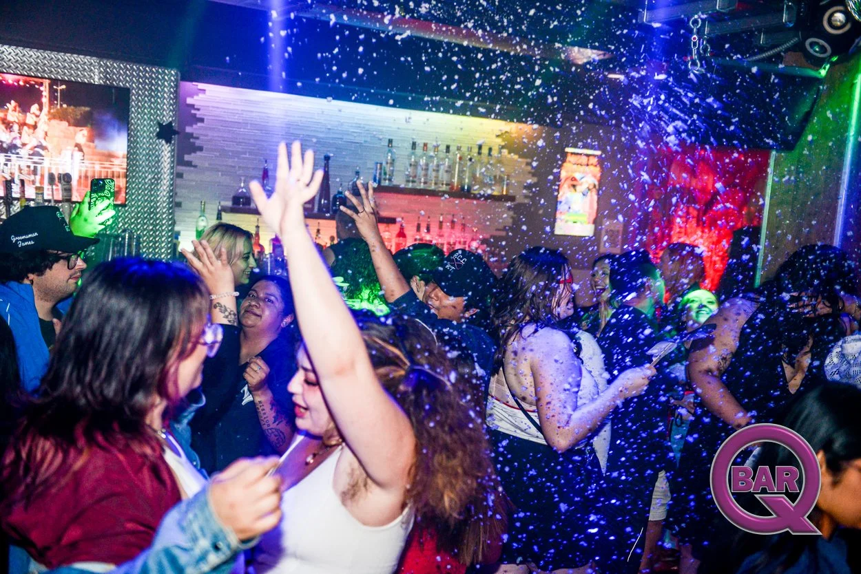 People dancing and socializing at a lively bar or nightclub, with colorful lighting and foam or snow falling from above.