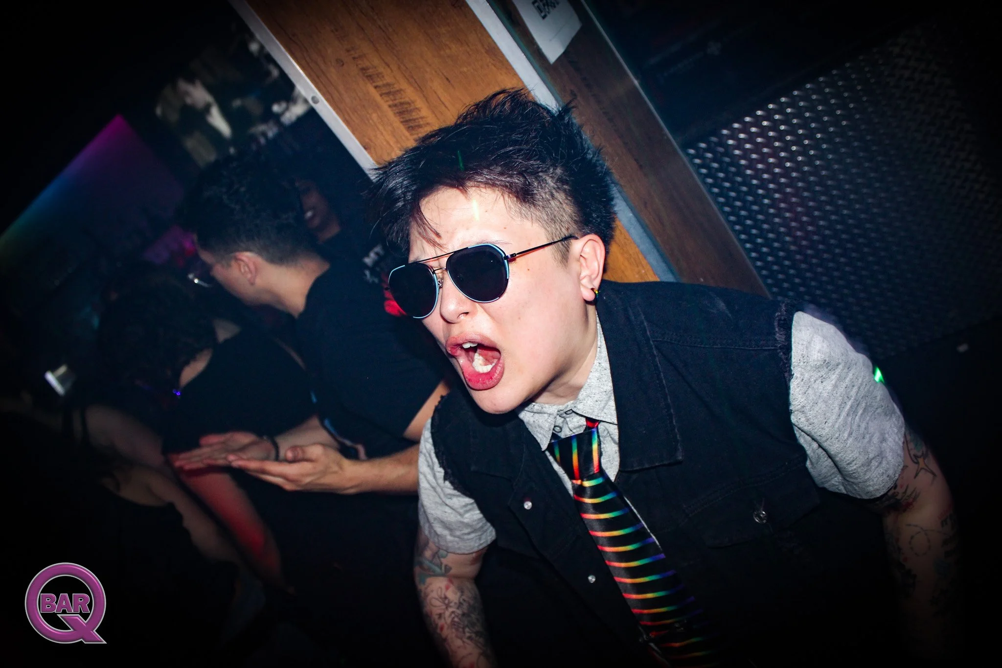 A person with short, spiky dark hair wearing sunglasses, a gray shirt, and a black vest, appears to be at a nightclub or bar with other people in the background, some with their heads bowed and hands clasped. The setting is dark with purple and pink 