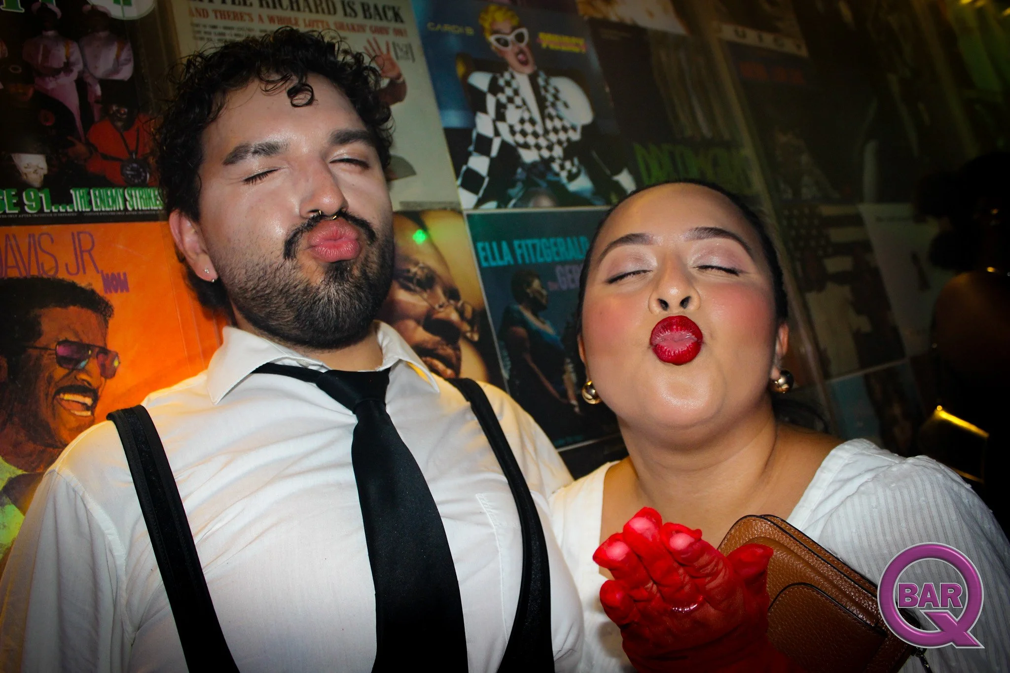 A man and woman with their eyes closed, standing close together. The man has curly black hair, a beard, wearing a white shirt with black suspenders and a black tie. The woman has makeup with bright red lipstick, and is holding a small brown purse and