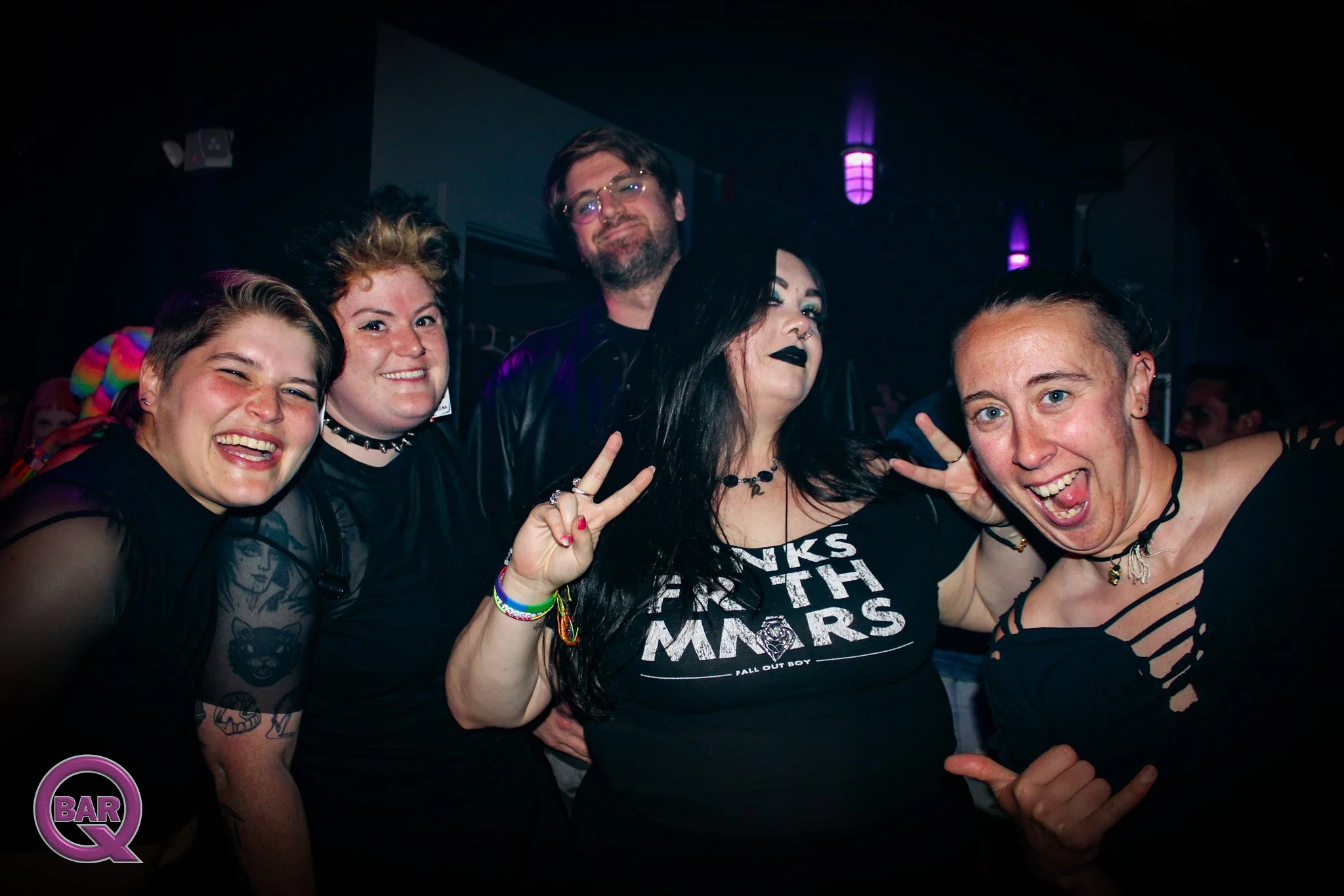 Group of five people at a bar or club, smiling and making peace signs, with dark background and purple lighting.