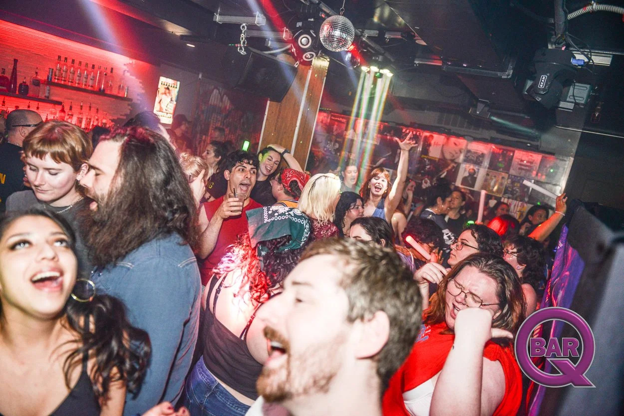 A crowded nightclub dance floor with people dancing and socializing. The scene is illuminated with colorful lights and a disco ball, creating a lively atmosphere. Bar decor is visible in the background.