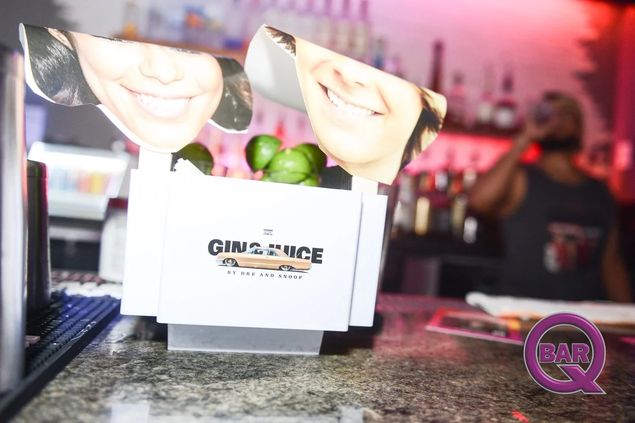Decorative bar sign featuring smiling adult faces with cutouts, a white box with the text 'GINGER JUICE by Dre and Snoop', and green limes inside the box, within a bar setting.