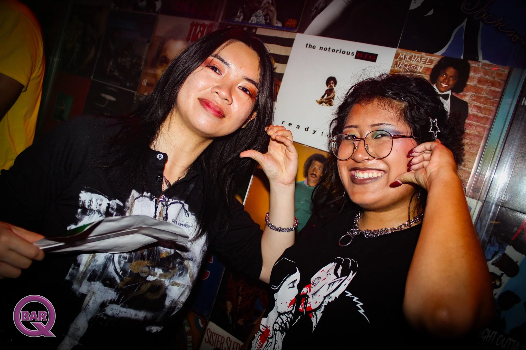 Two women smiling and posing together at a gathering, with posters of Michael Jackson and other music artists in the background, and a purple "BAR Q" logo at the bottom left corner.
