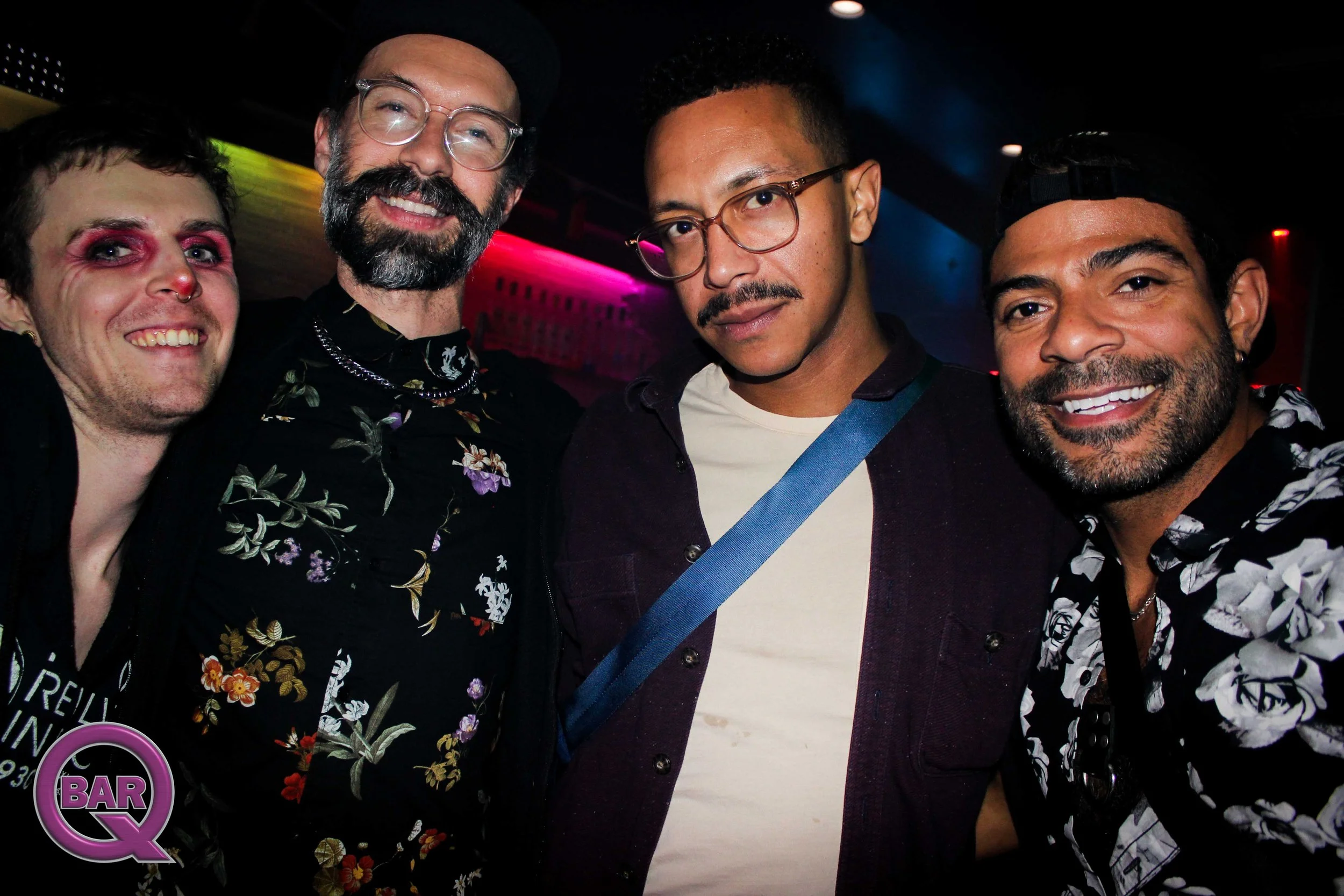 Four men standing close together at a bar or nightclub, smiling at the camera. The background is dark with colorful lighting.