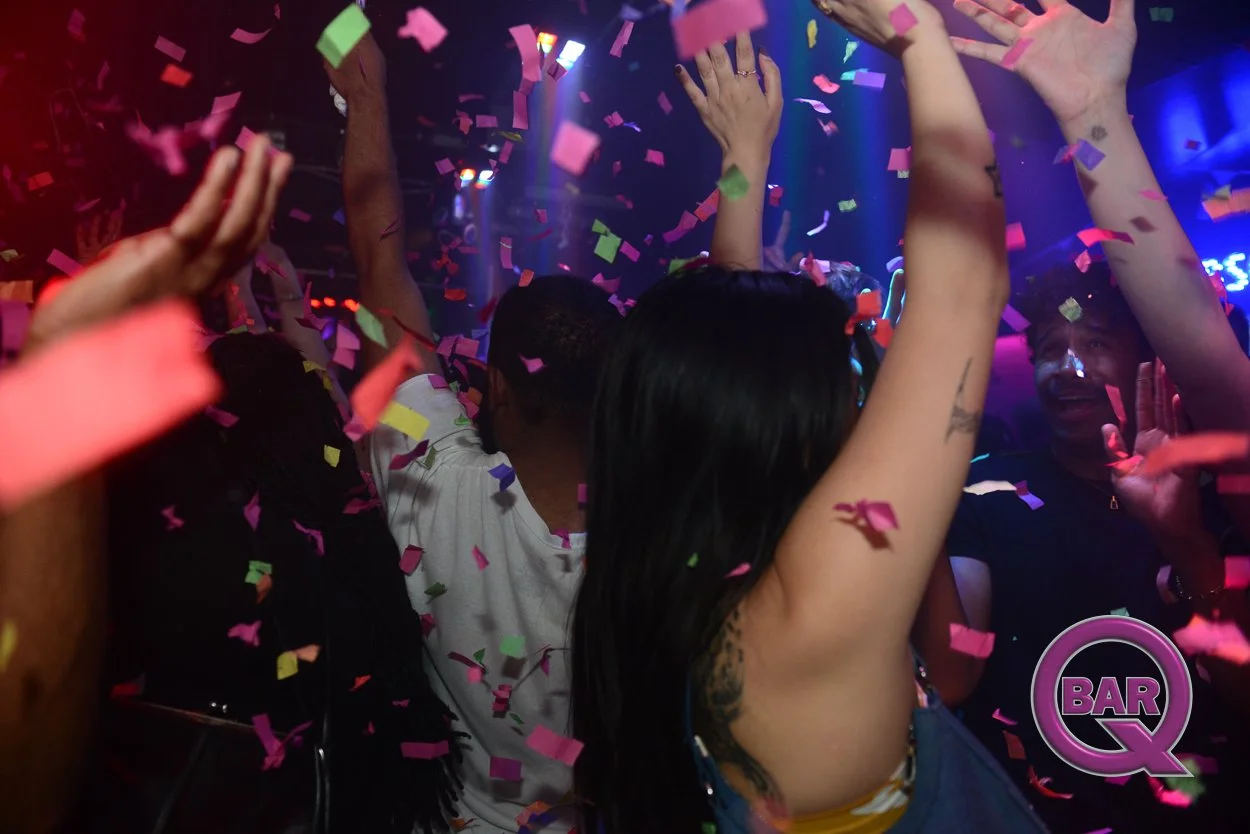 People dancing and celebrating in a nightclub with colorful confetti falling, neon lighting, and a bar logo visible in the bottom right corner.