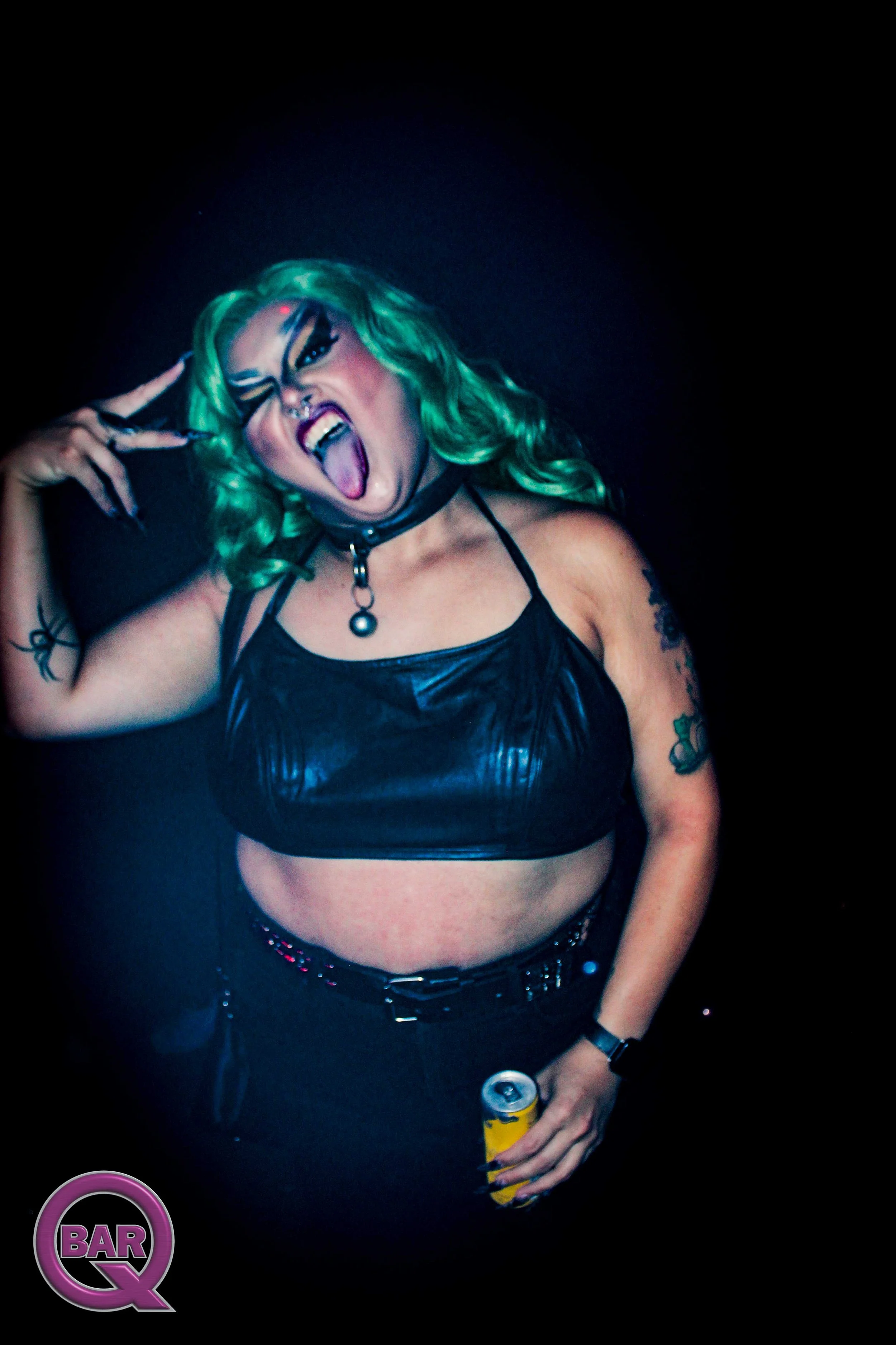Person with bright green hair, heavy makeup, tongue out, making a rock sign with hand, wearing black leather crop top, tattoos, holding a yellow can, dark background with a 'Bar Q' logo