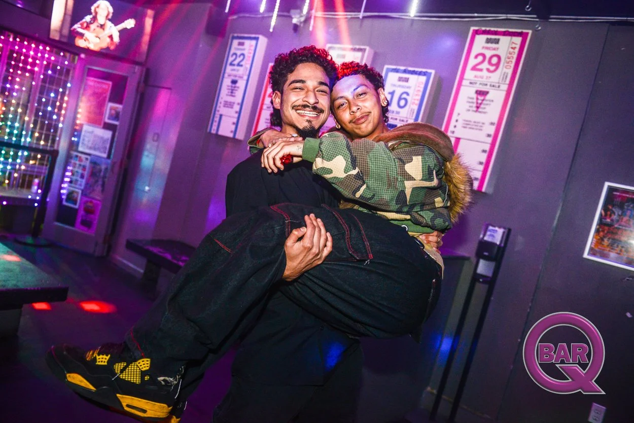 Two young men smiling, one carrying the other in a nightclub or bar with neon lights and posters on the wall, including a bottle of water and a 'Bar Q' logo.