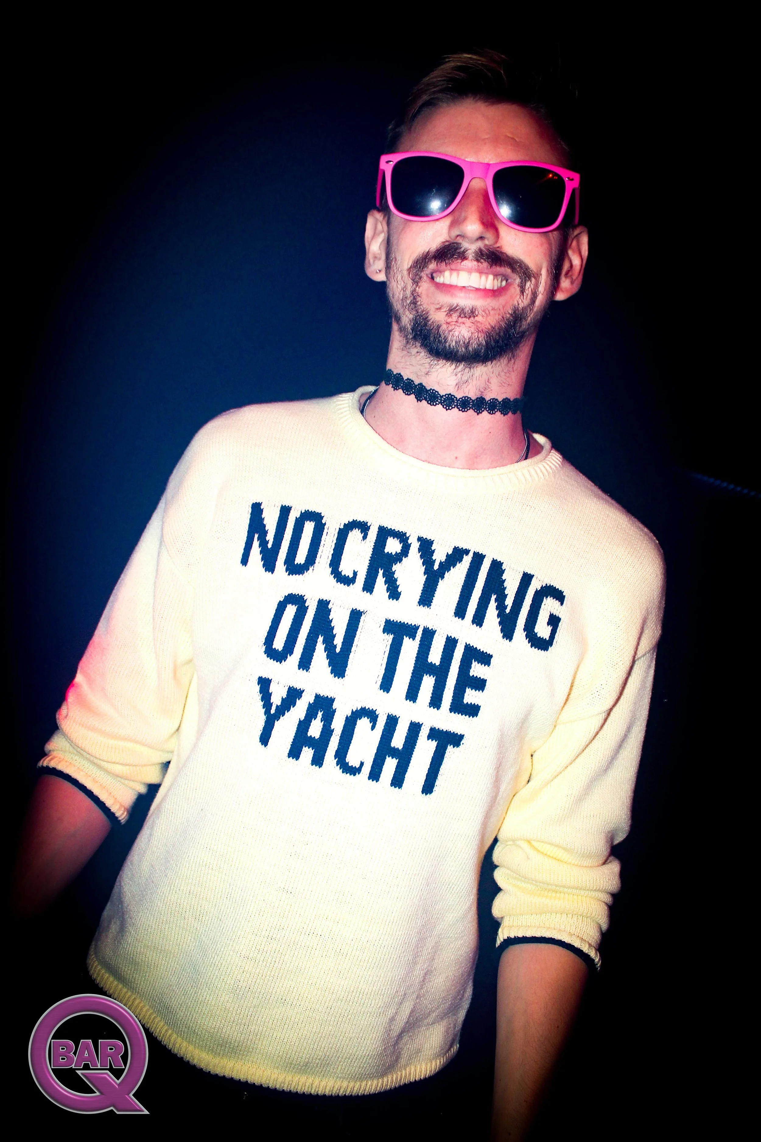 A person smiling, wearing pink sunglasses, a choker, and a yellow sweater with the text 'NOCAYING ON THE YACHT.' The background is dark, and there is a 'BAR Q' logo in purple and black in the bottom left corner.