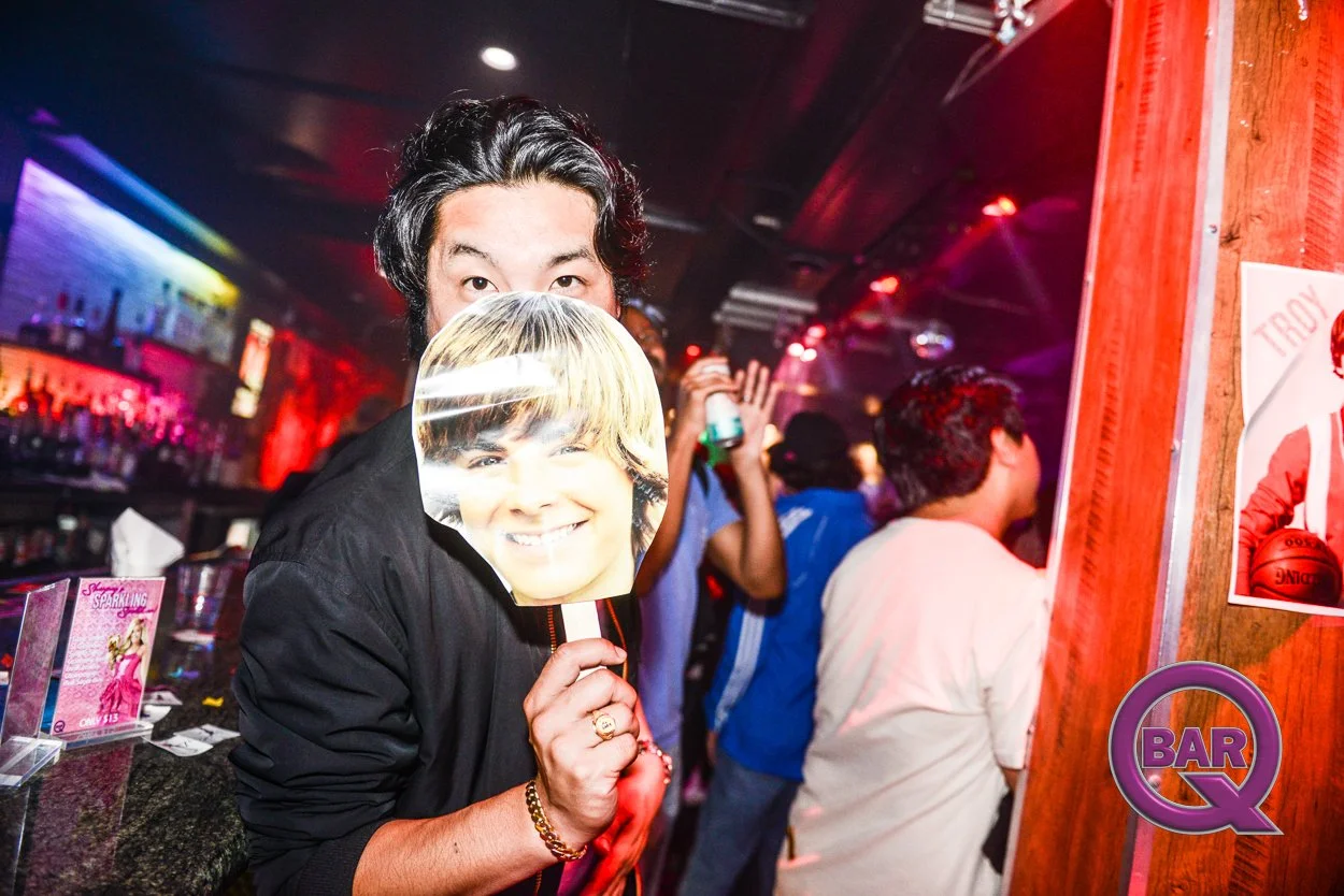 A man at a bar party holding a mask with a smiling woman's face, surrounded by other people, neon lights, and bar decor.