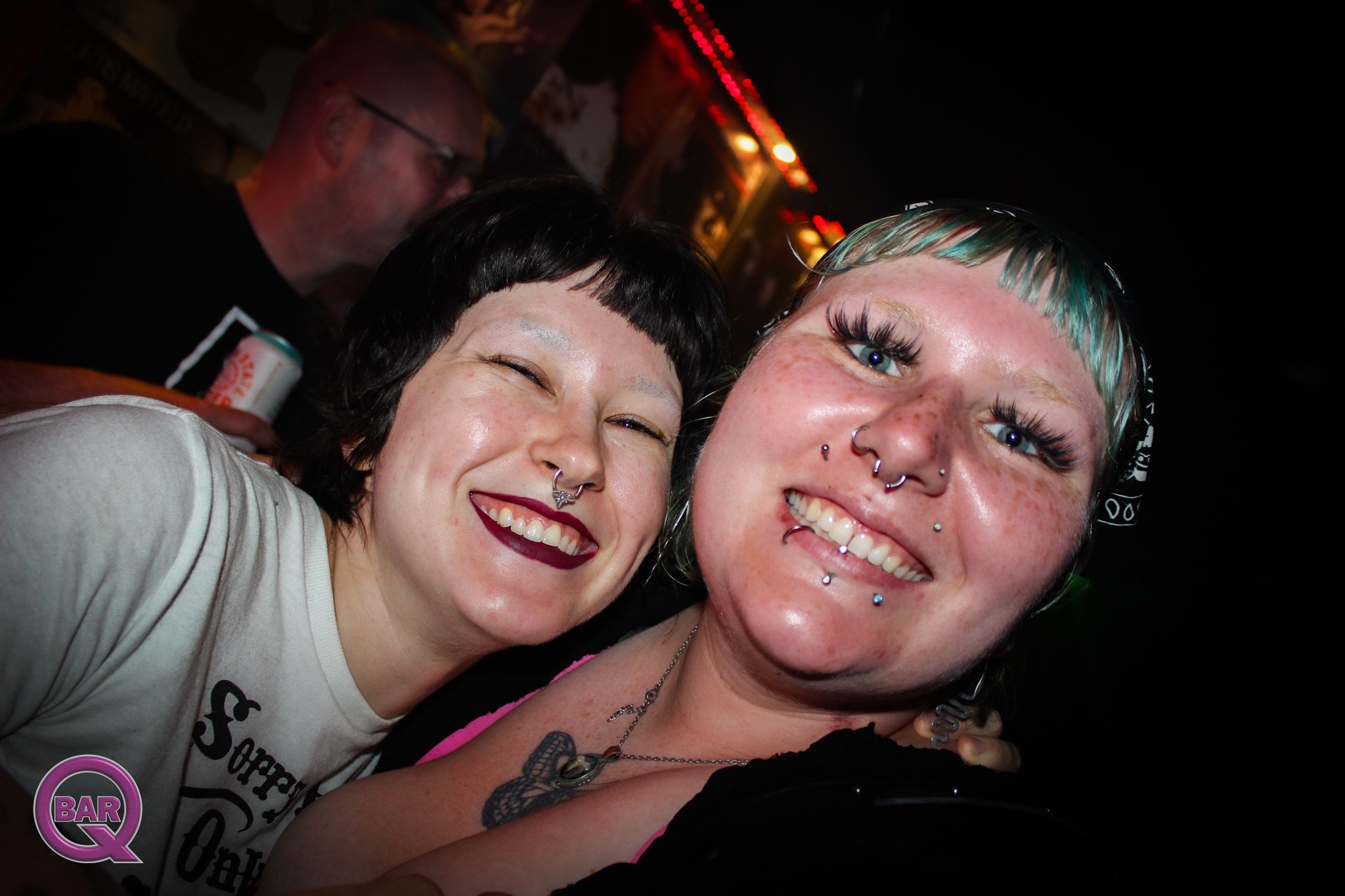 Two women taking a selfie at a bar, smiling, with tattoos and piercings, in a dimly lit setting with red lights in the background.