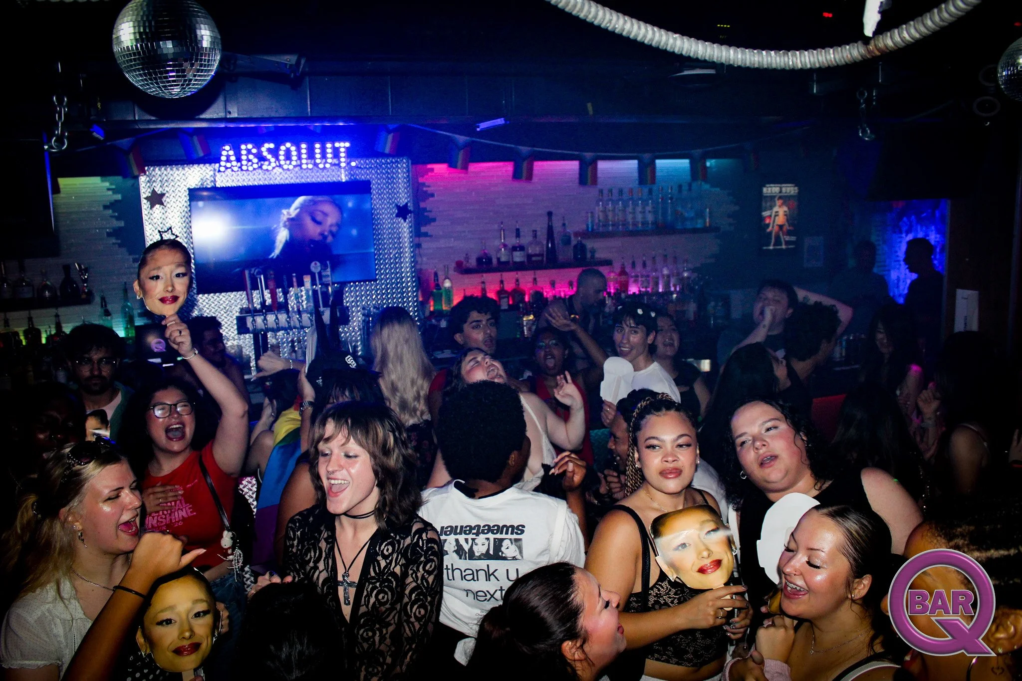 Crowd of diverse people enjoying a lively party at a bar with colorful lights and a large screen displaying a woman. Some people are wearing masks with faces on them, and a disco ball hangs from the ceiling.