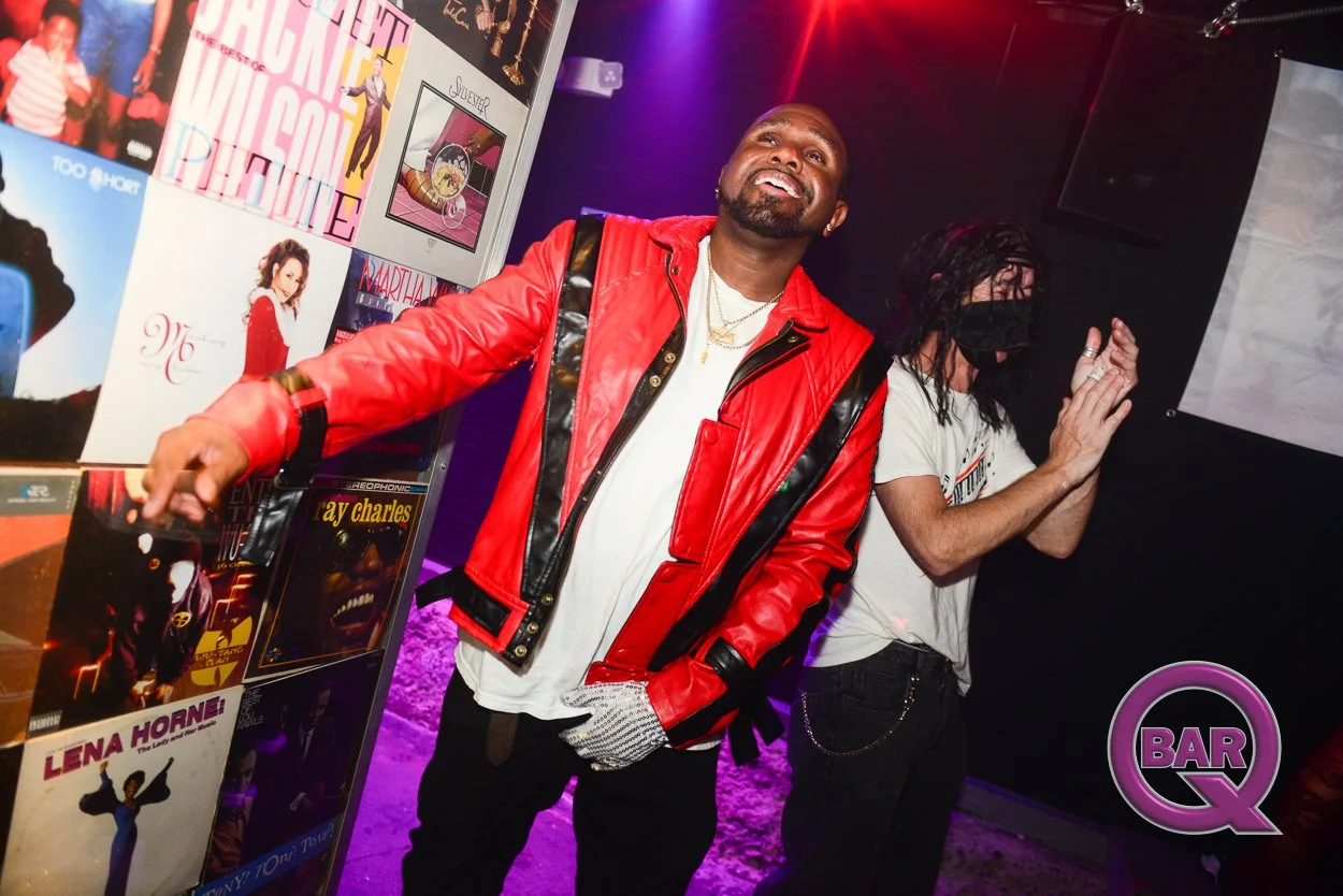 Two men at a music event, one wearing a red leather jacket and the other wearing a mask and a white t-shirt, standing next to a wall with various album covers displayed, including Lena Horne and Ray Charles, in a nightclub or bar setting with purple 