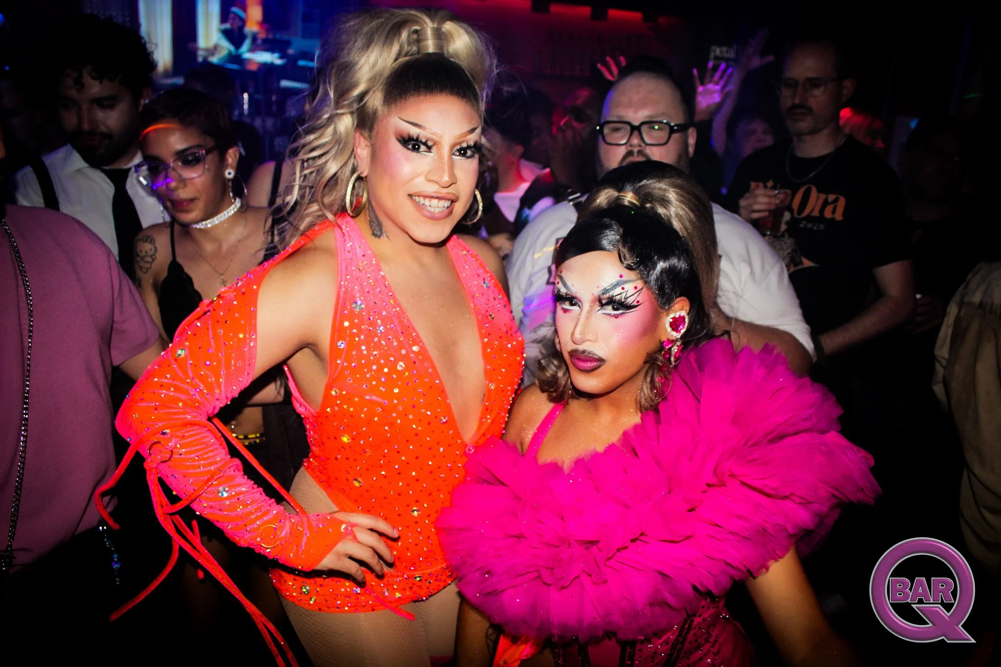 Two people in vibrant outfits posing at a party, with a crowd in the background on a dance floor.